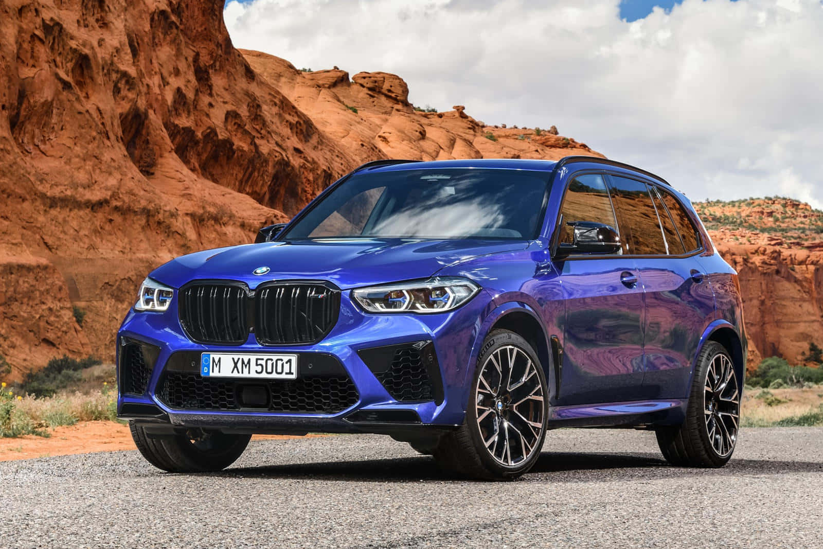優雅BMW X5於壯麗風景中 壁紙 — 一架鮮藍色BMW XM SUV停泊在碎石上，背景有紅色岩層和陰天。