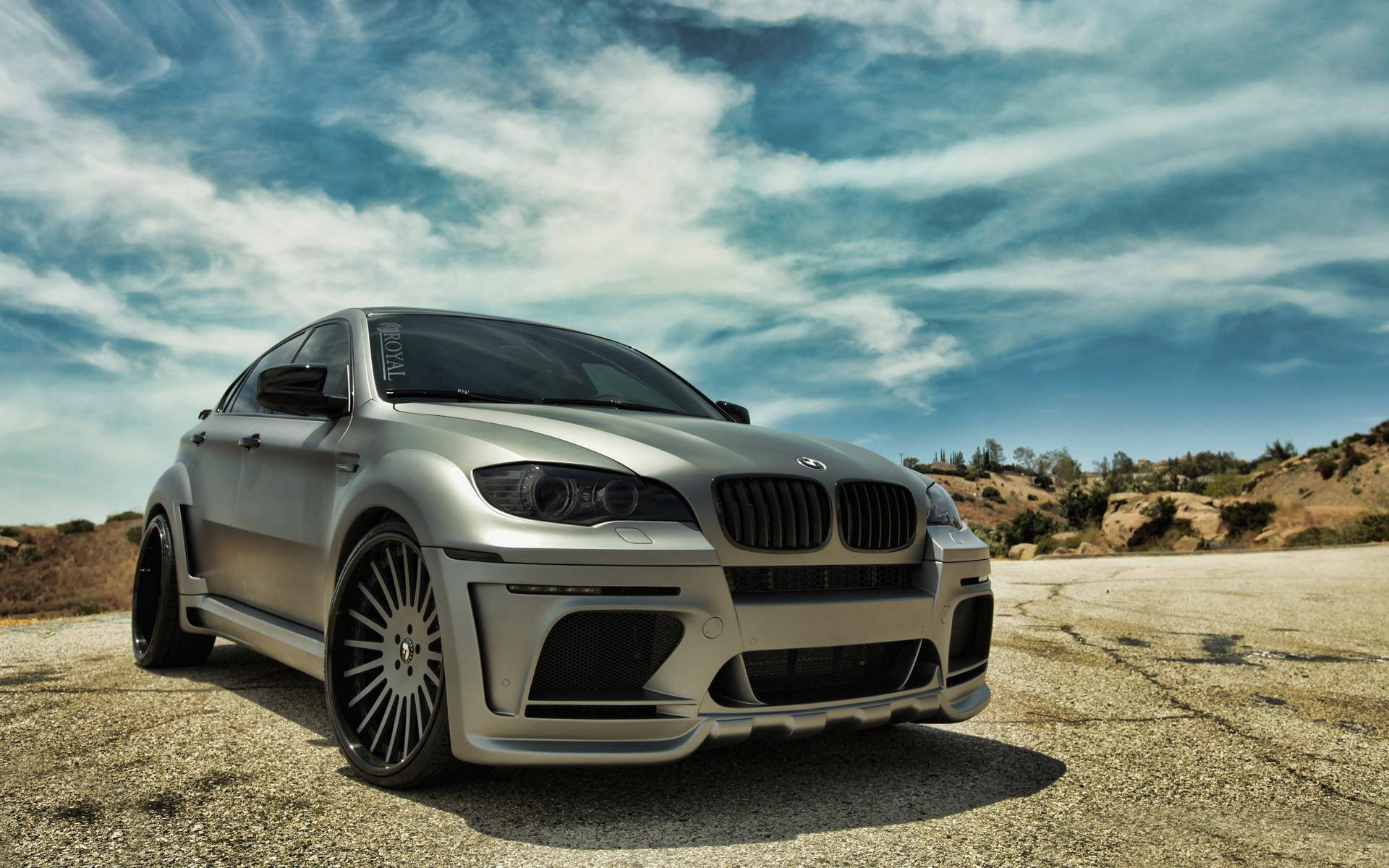 BMW X6 M 夢幻雲朵 壁紙 — 一輛銀色BMW M6雙門轎跑車，配備寬體套件和定制輪圈，停在裂開的瀝青路面上，天空烏雲密佈，氣氛戲劇化。