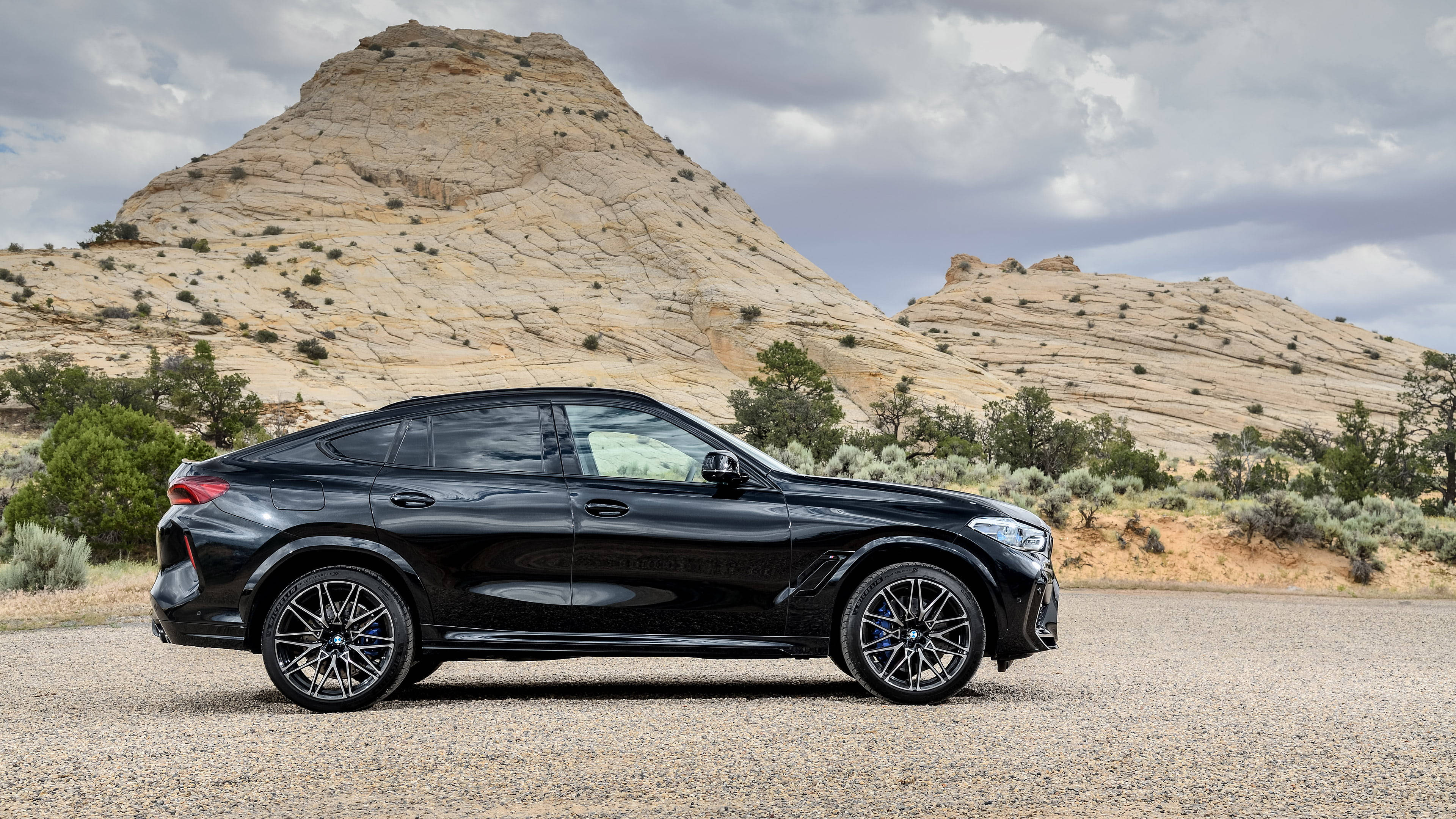 BMW X6 M 沙漠道路 壁紙 — 一架時尚黑色BMW X6 SUV停泊在碎石上，背景是崎嶇的岩石山丘和陰天。