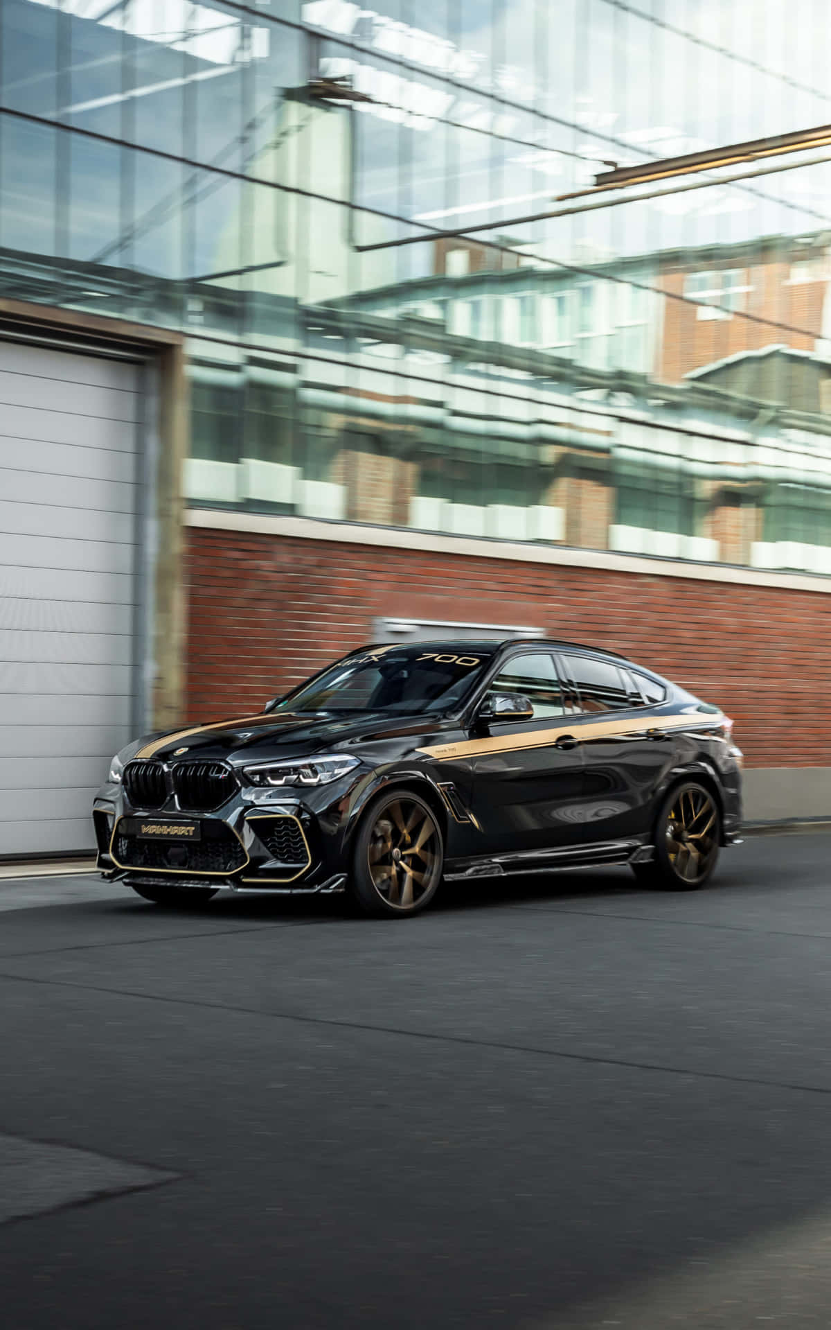 BMW X6 M 的驚豔動力 壁紙 — 一輛帶金色裝飾的時尚黑色BMW X6 M700在城市的街道上飛馳而過，經過一座現代化的玻璃和磚砌建築。