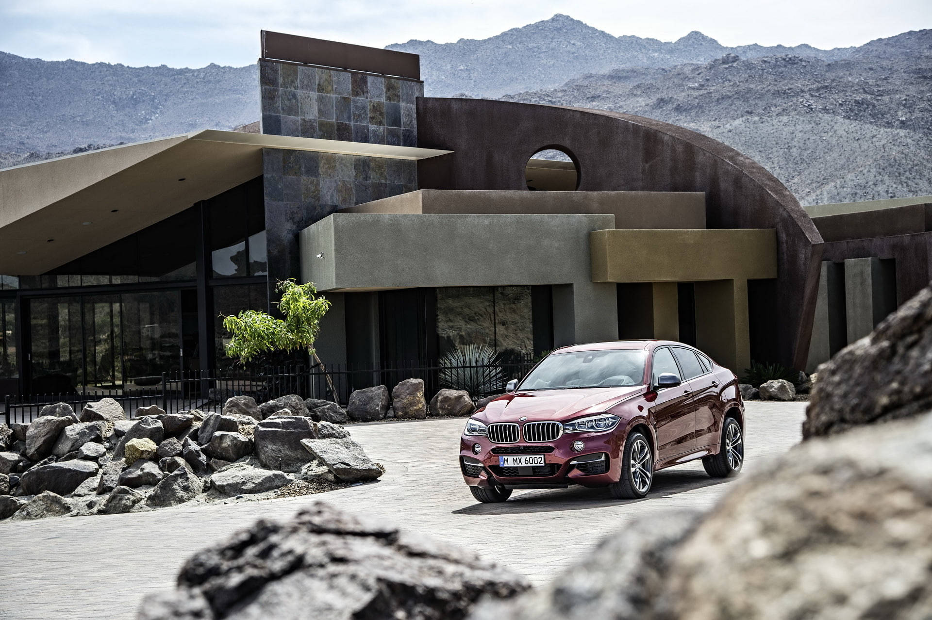 BMW X6 M 與石屋 壁紙 — 一架栗色BMW X6停泊在現代沙漠屋前，背景有山脈。