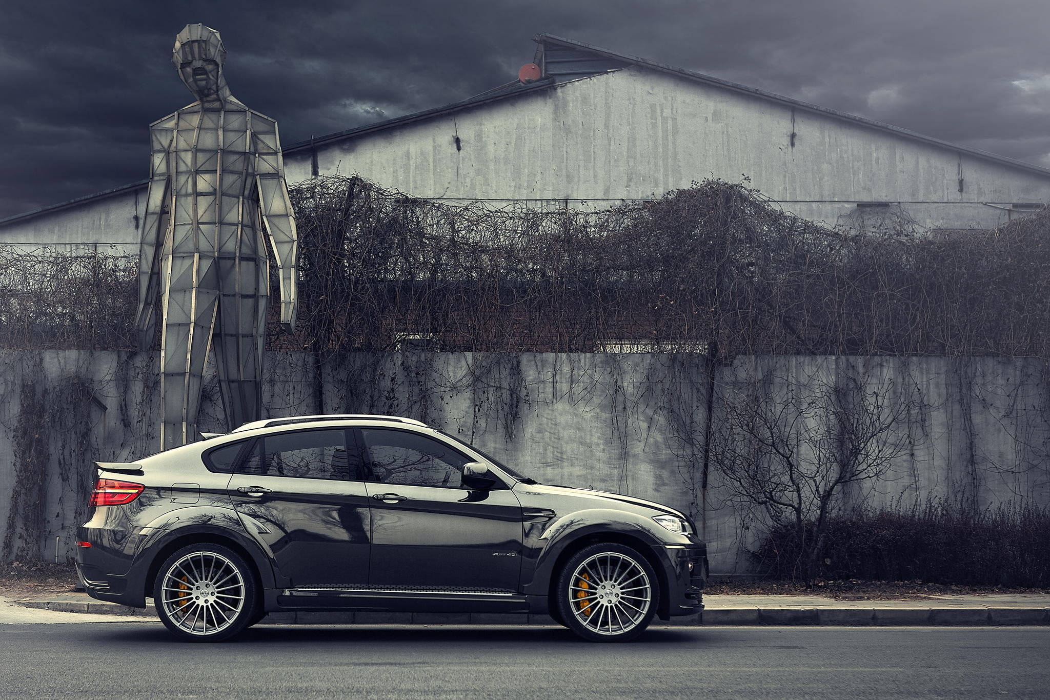 BMW X6 M 與雕塑 壁紙 — 一架時尚的黑色BMW X6停在一座高聳的幾何人形雕像旁，天空陰沉多雲。