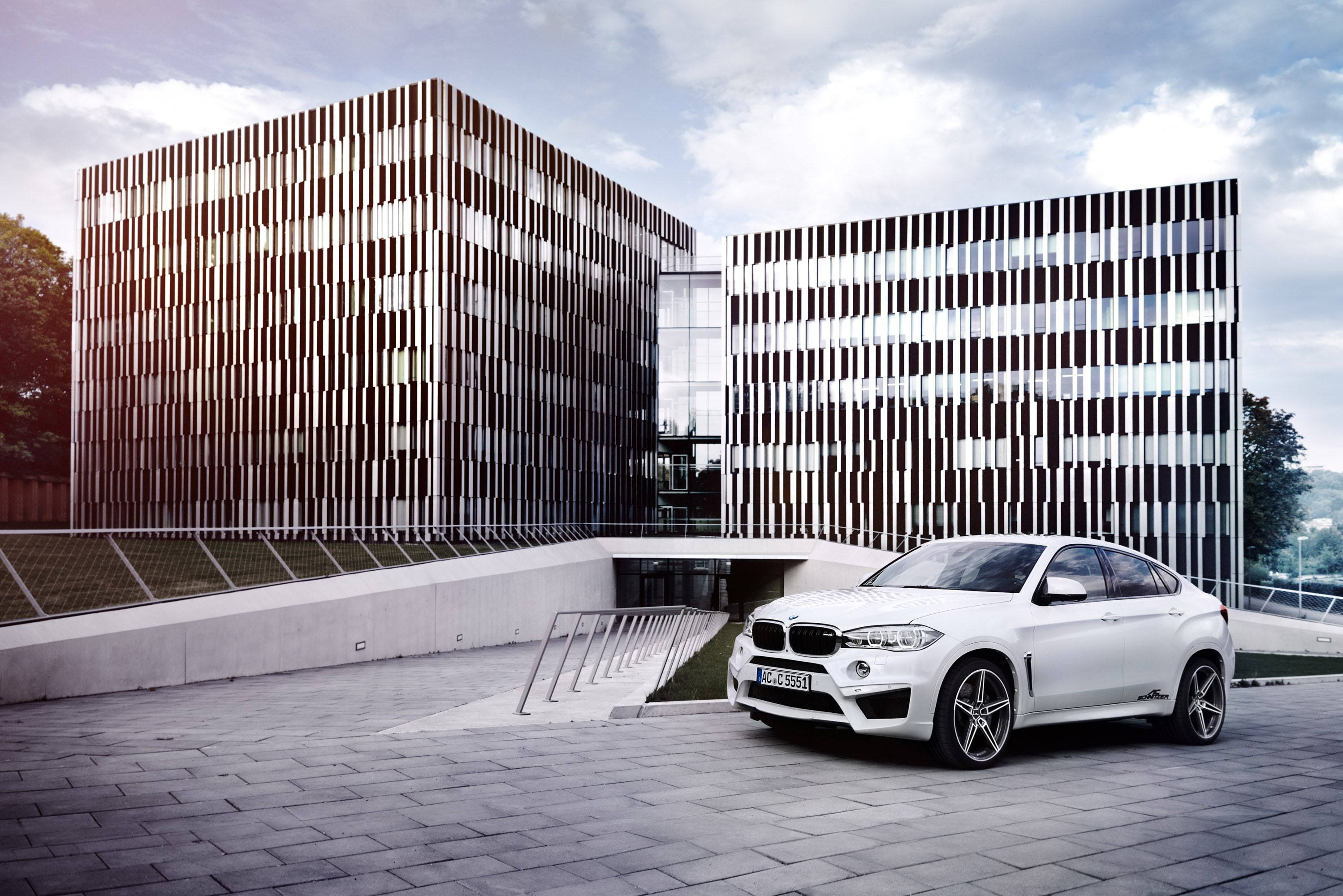 BMW X6 M停泊於德國ICT立方體建築前 壁紙 — 一輛時尚的白色BMW X6停泊在鋪砌的廣場上，前方是現代建築，其立面帶有垂直板條，天空部分多雲。