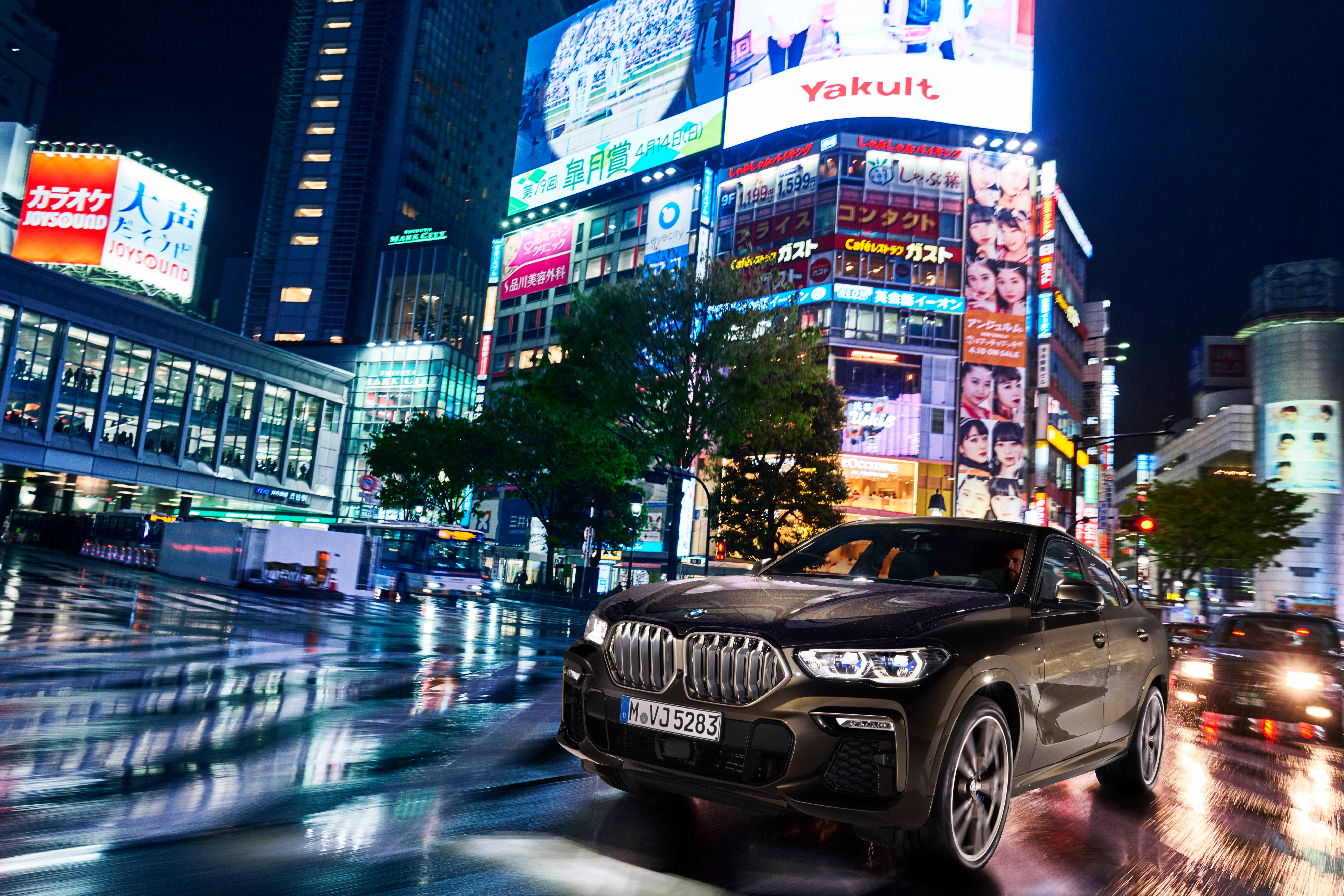 BMW X6 M夜景城市燈光 壁紙 — 一輛時尚的黑色BMW X6在雨濕的東京夜街上飛馳，四周是耀眼的霓虹燈招牌和發光的摩天大樓。