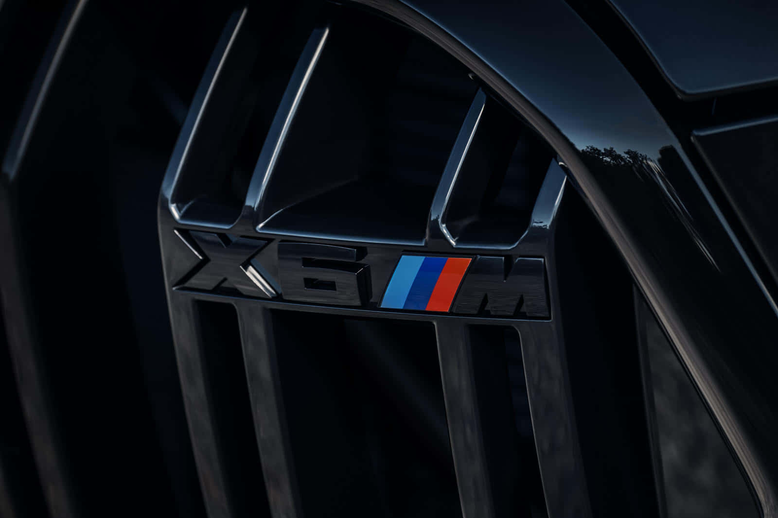 BMW X6 M是真正的駕馭巔峰 壁紙 — 近距離拍攝黑色BMW X6M鬼面罩徽章，帶有藍、紅、紫條紋，在戲劇性燈光下呈現。