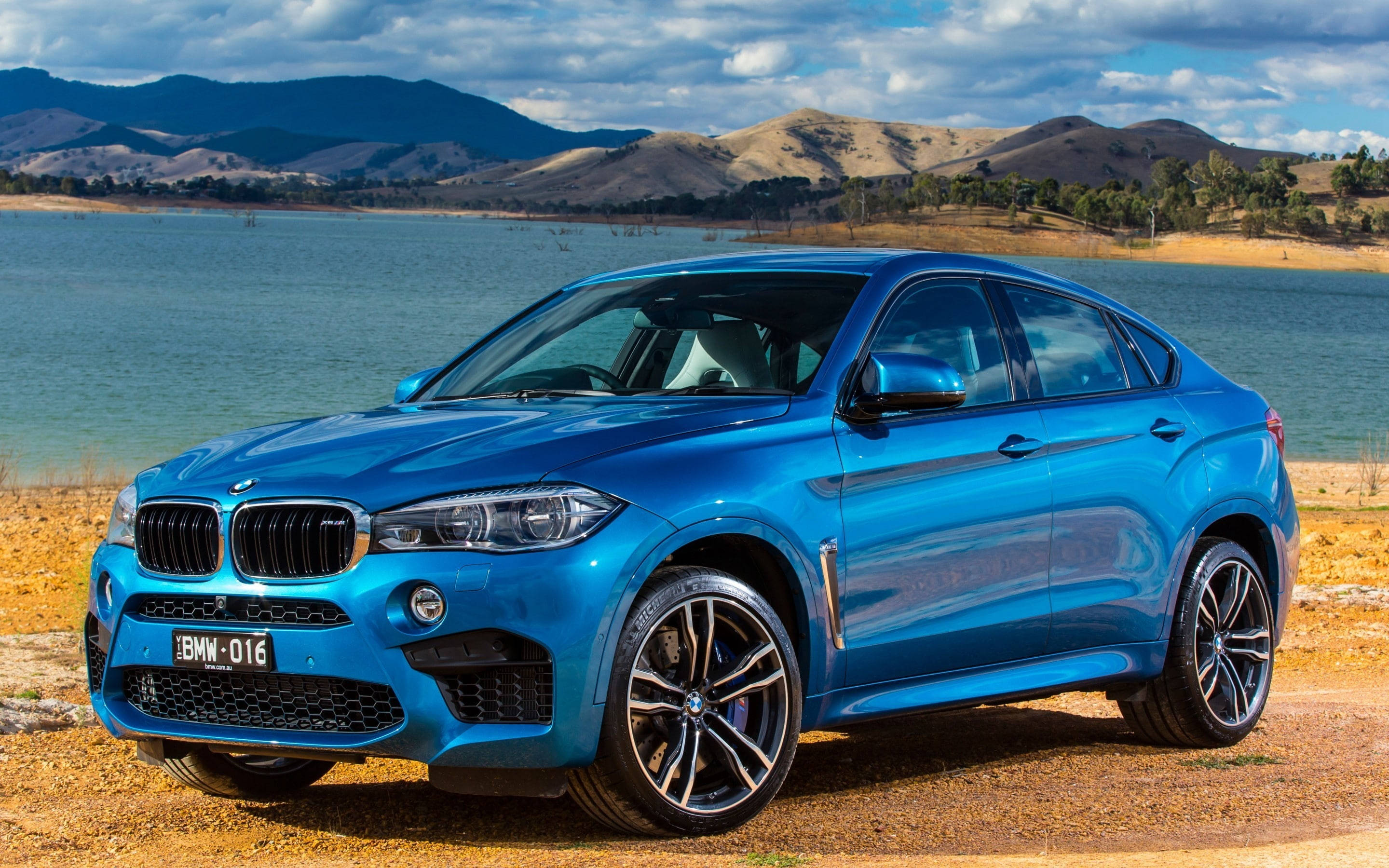 BMW X6 M海景風光 壁紙 — 一輛鮮藍色的BMW X6 M停在平靜湖泊旁的碎石上，背景有起伏的山丘和多雲的天空。