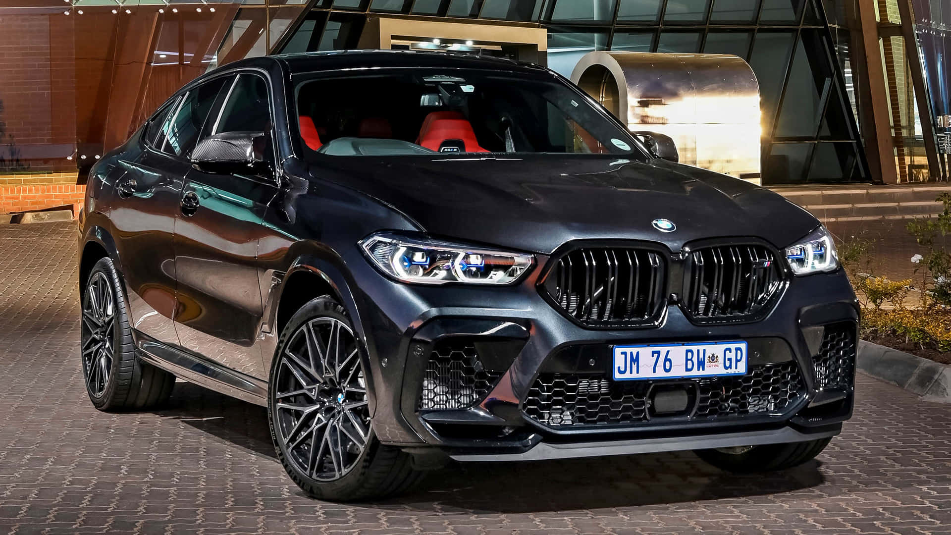 BMW X6 M的強勁引擎 壁紙 — 一輛時尚的黑色BMW X6 M在夜間停放在圖案人行道上，背景是現代建築。