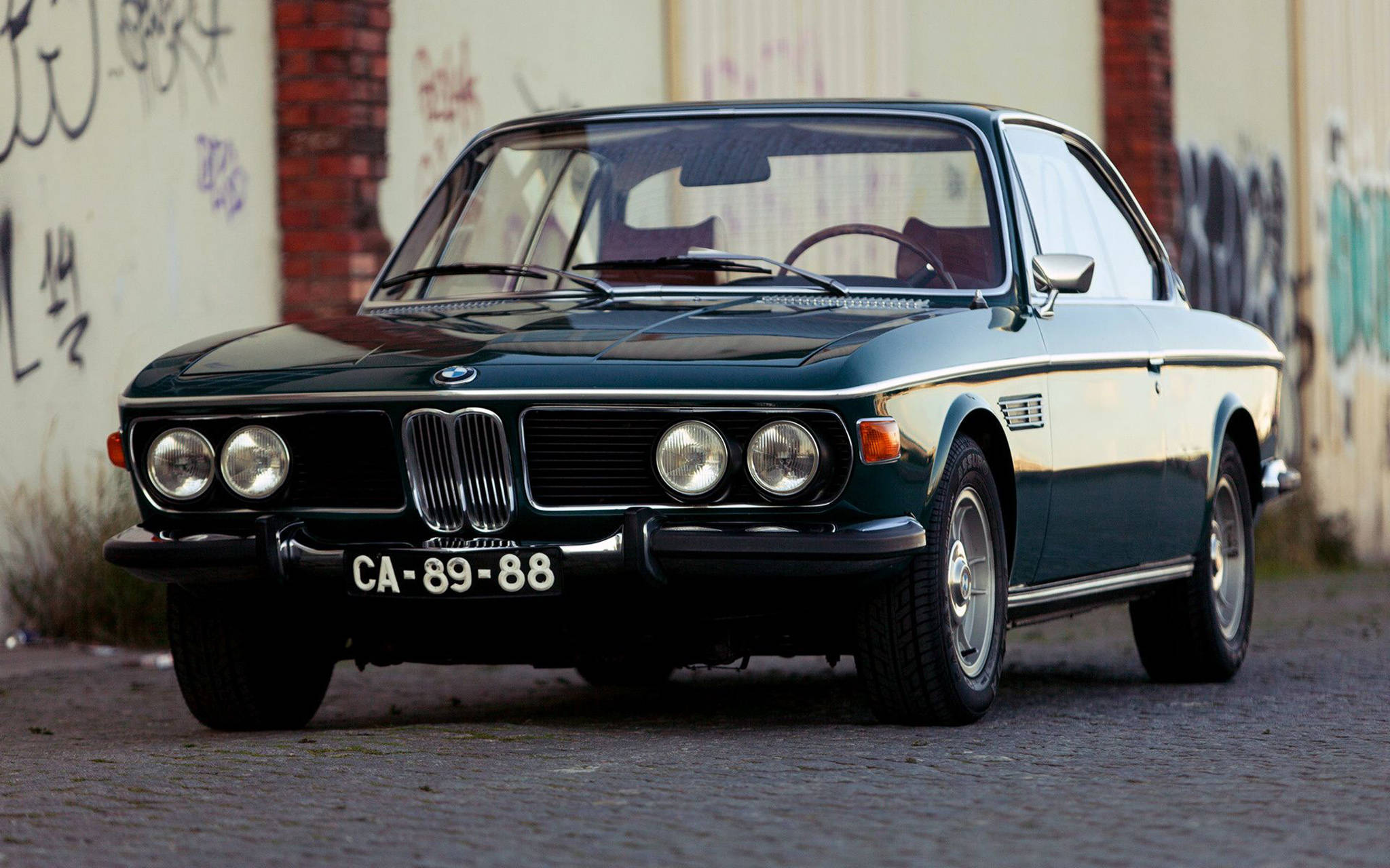 金屬藍經典BMW 壁紙 — 一架復古深綠色寶馬6系轎跑車停泊在鵝卵石街道上，旁邊是佈滿塗鴉的牆壁。