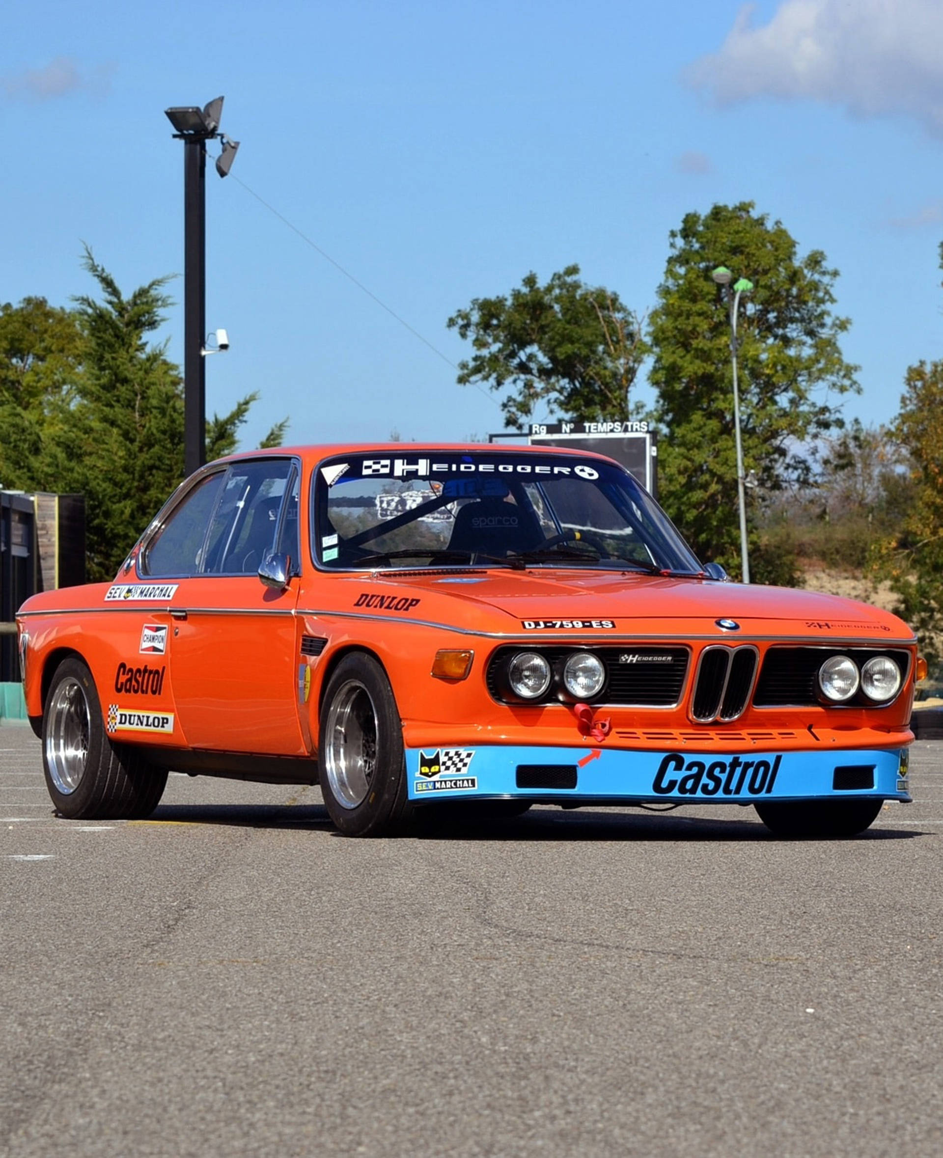 深橙色經典BMW 壁紙 — 一輛充滿活力的橙色復古賽車，帶有藍色點綴和Castrol貼紙，停在晴朗藍天下的瀝青路面上。