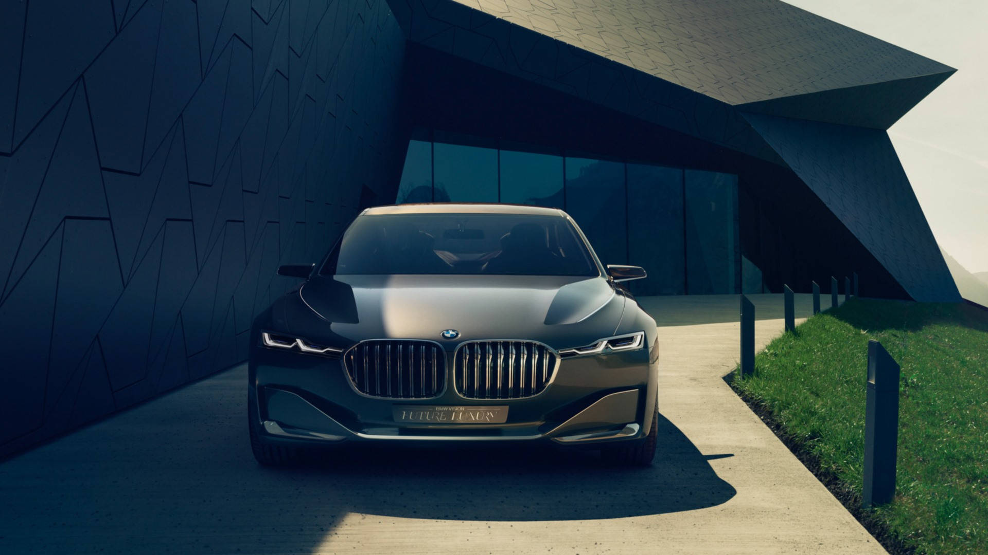 BMW 豪華汽車待命 壁紙 — 一輛時尚的深灰色BMW豪華轎車停放在現代幾何牆壁建築和混凝土路旁綠草地的現代棱角建築下。