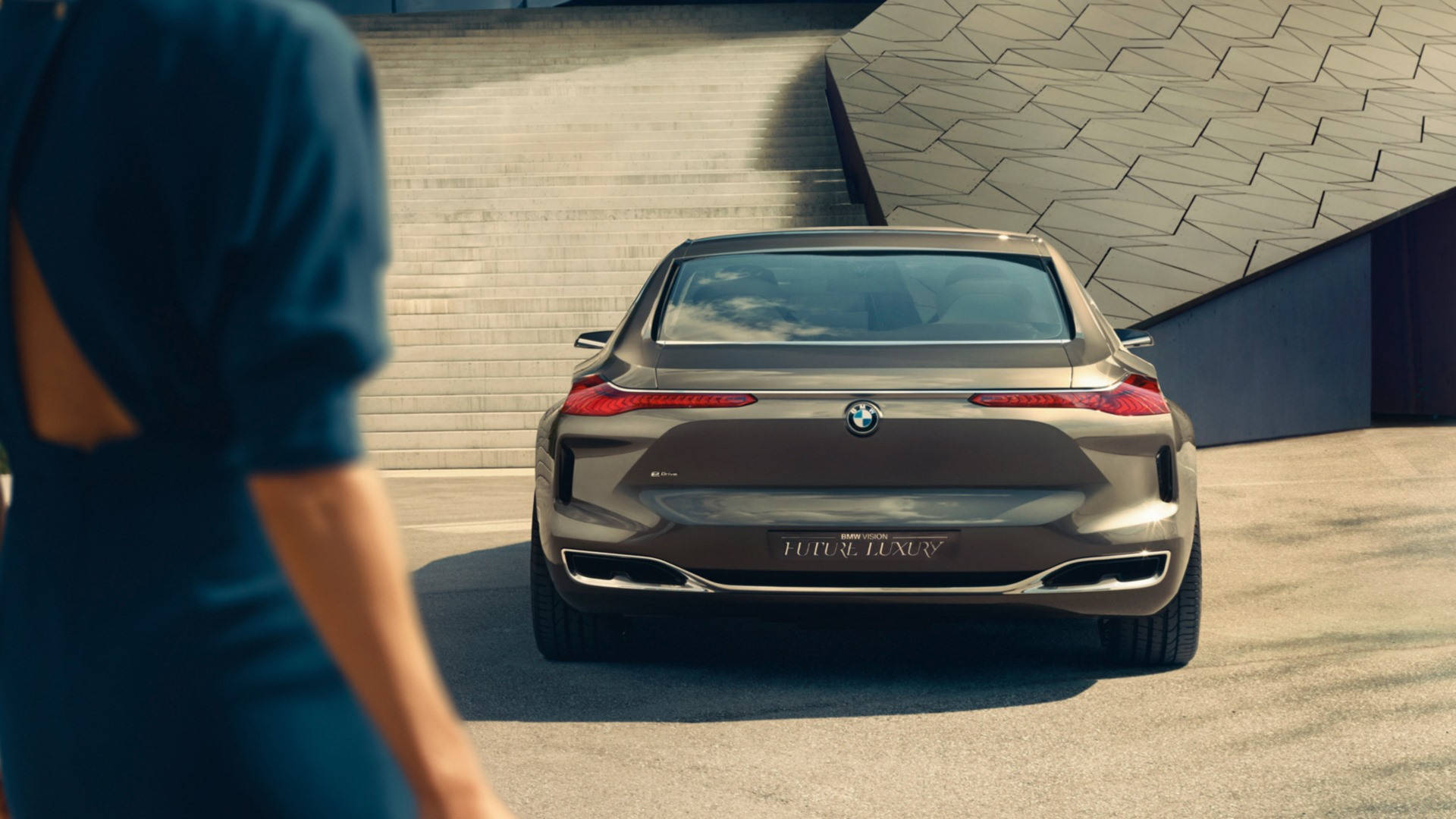 BMW 豪華汽車後視 壁紙 — 一輛流線型的青銅色BMW概念車停放在現代幾何建築前，一位身穿藍色連衣裙的女性走過。