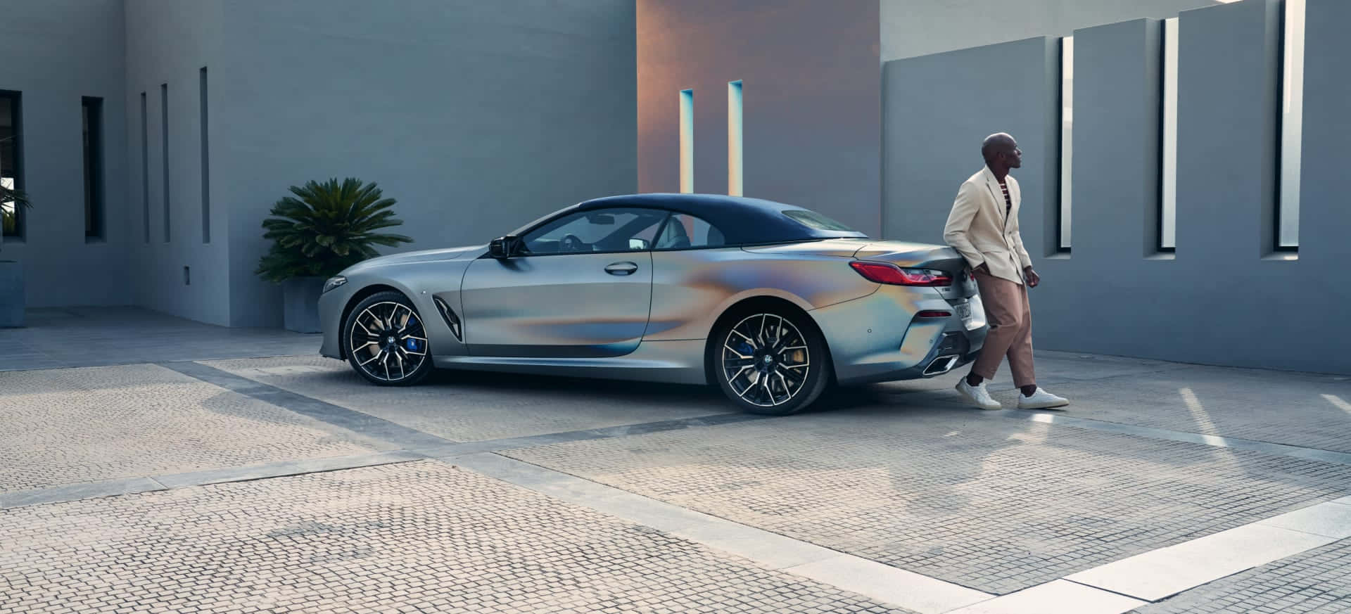 BMW的奢華與創新 壁紙 — 一名身穿米色西裝的男子走過一輛停在現代建築風格圖案庭院中的時尚銀色敞篷車。