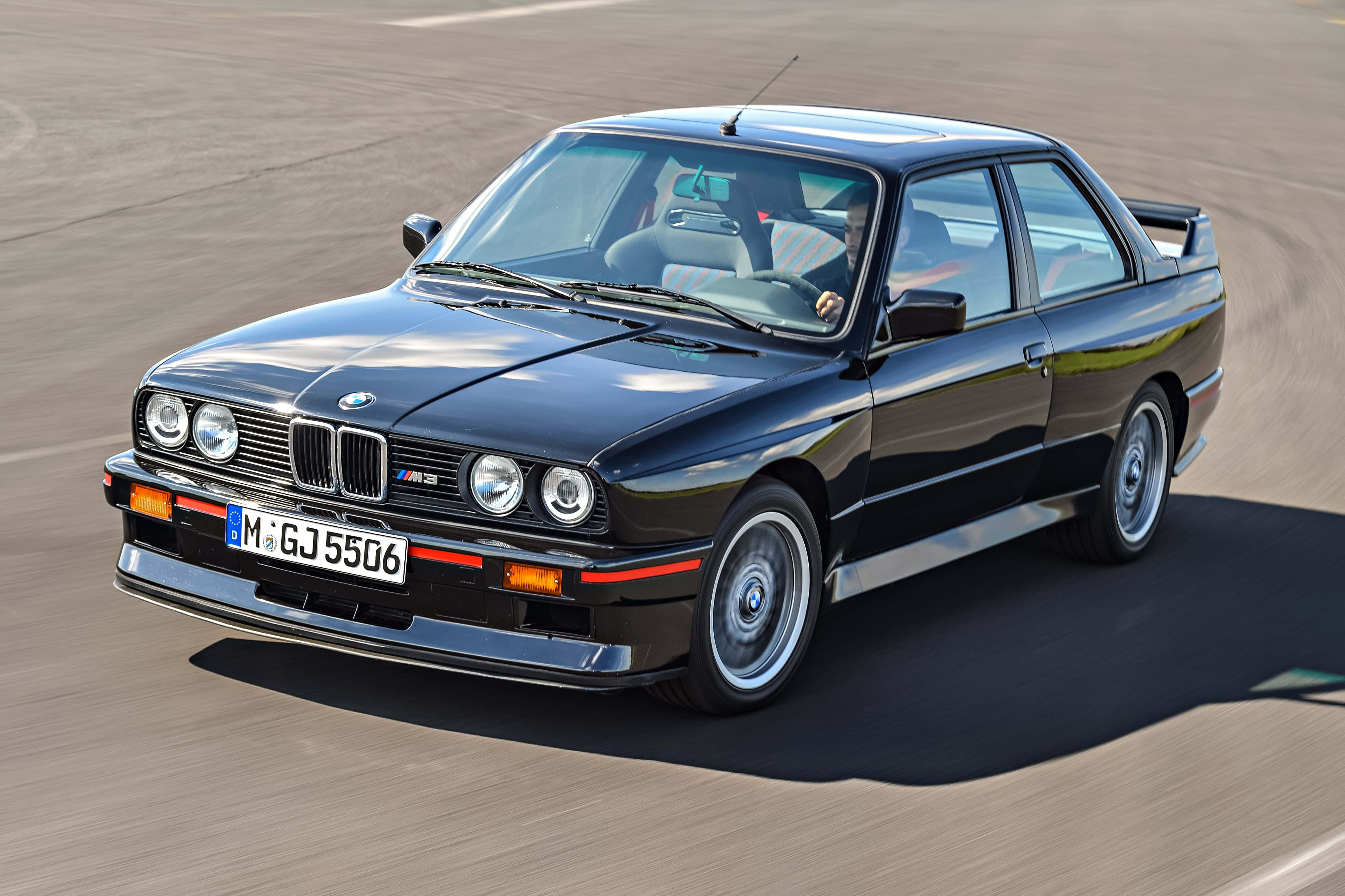 BMW車款於樹影剪影背景中顯得格外突出 壁紙 — 一架流線型黑色BMW M3雙門跑車在陽光照耀的賽道上疾馳，動態模糊效果凸顯其強勁性能。