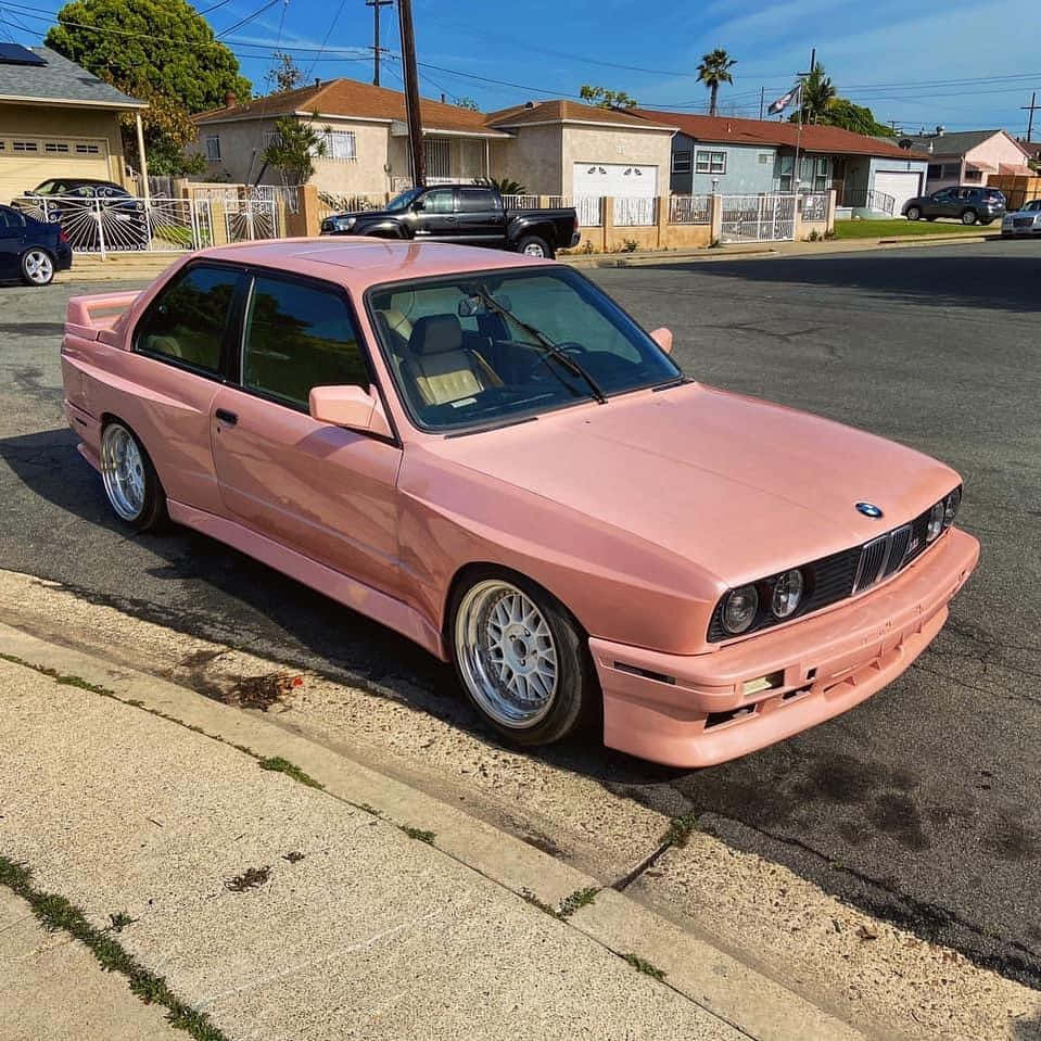BMW車款駛過高速公路，夕陽映照 壁紙 — 一架光面粉紅色BMW E30雙門跑車停泊在陽光明媚的郊區街道上，背景有棕櫚樹和房屋。