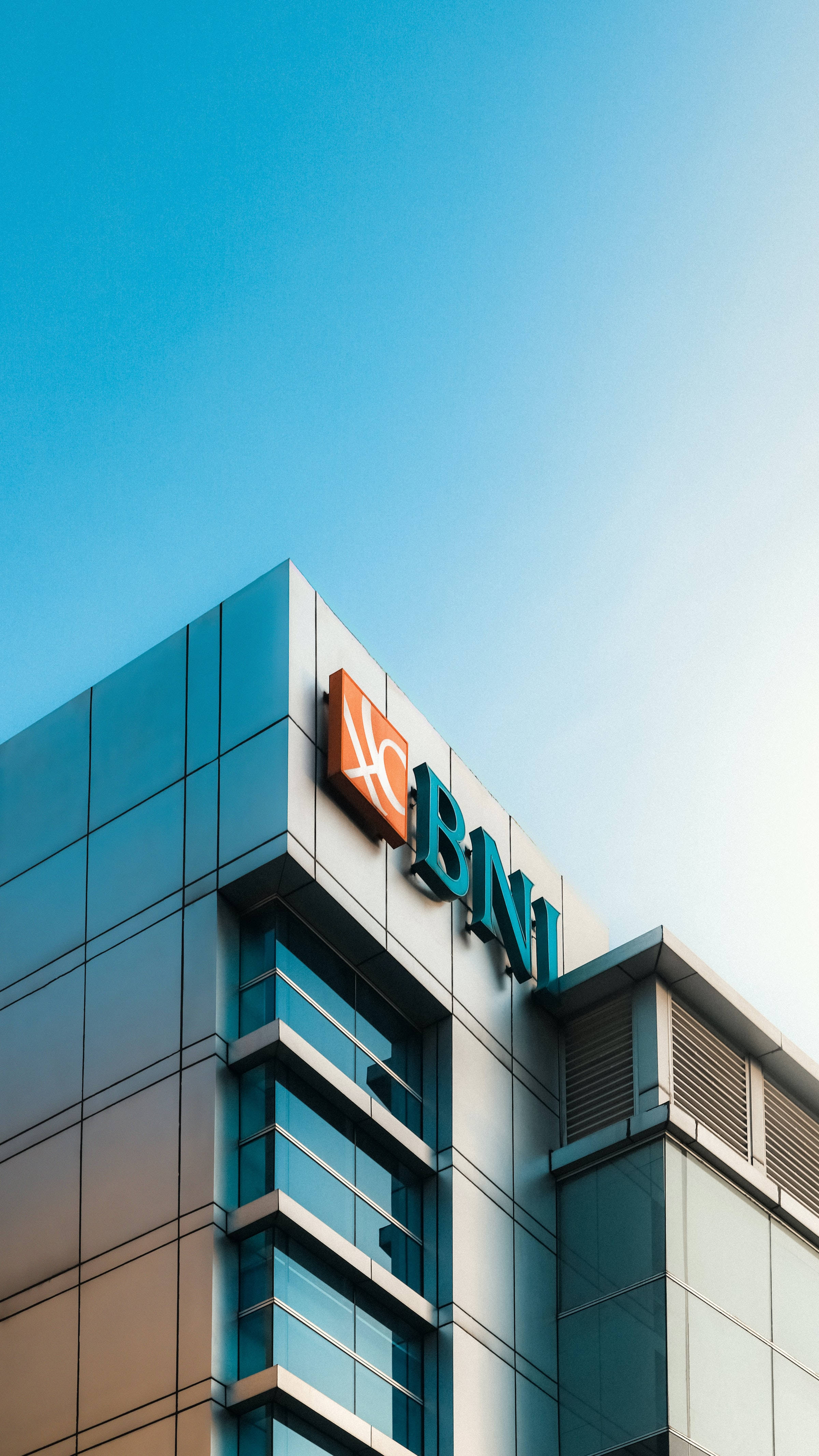 印尼BNI Syariah銀行外觀 壁紙 — 白天晴朗藍天下帶有BNI標誌的現代玻璃建築。