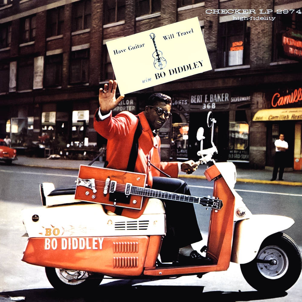 Bo Diddley騎橙色摩托車 壁紙 — Bo Diddley 微笑著騎著橙色滑板車，背著結他，手持寫有「Have Guitar Will Travel」的牌子。