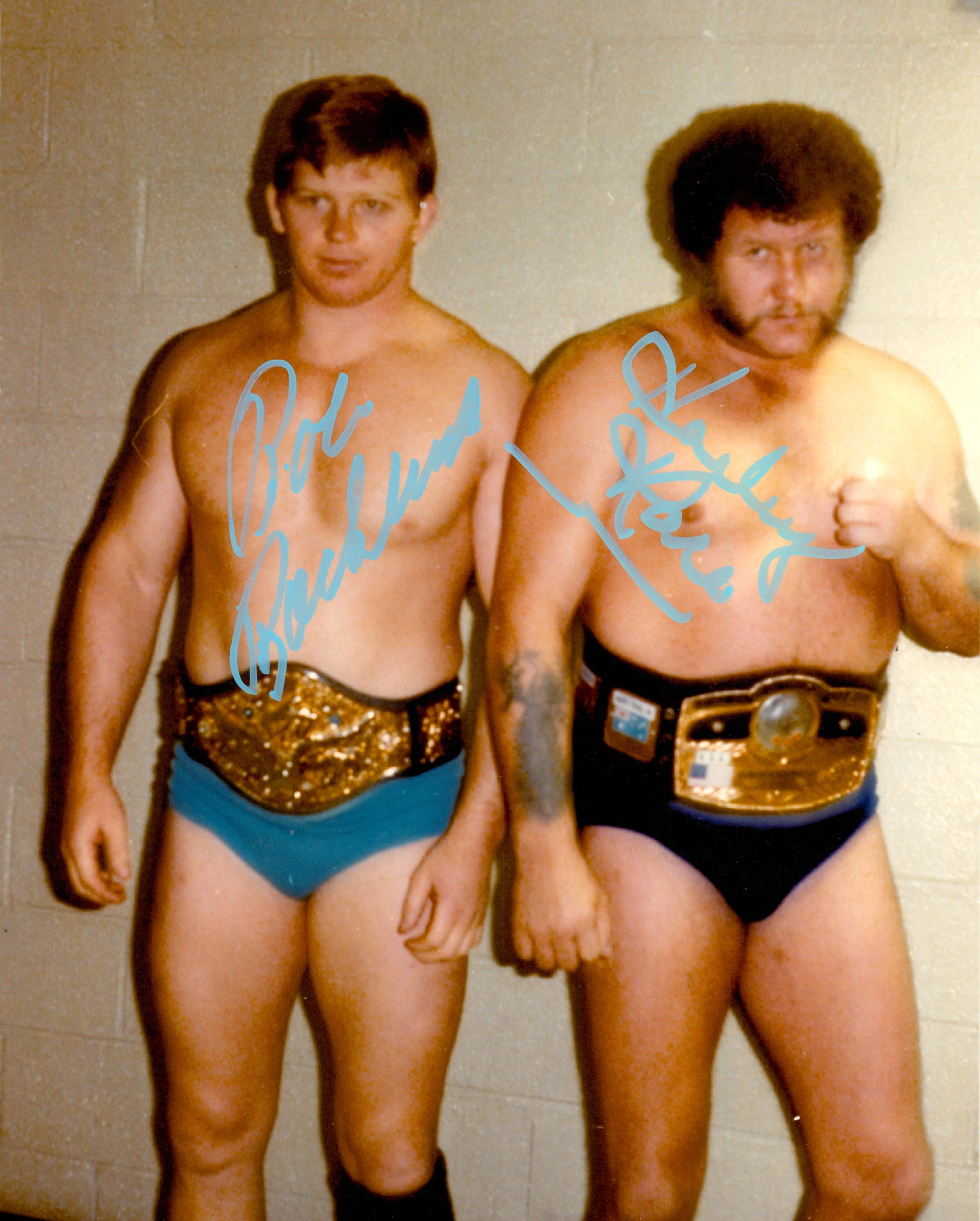Bob Backlund 與 Harley Race 雙人簽名壁紙 壁紙 — 兩名肌肉發達的摔角手穿著藍色短褲和冠軍腰帶，自信地靠在普通的煤渣磚牆上擺姿勢。