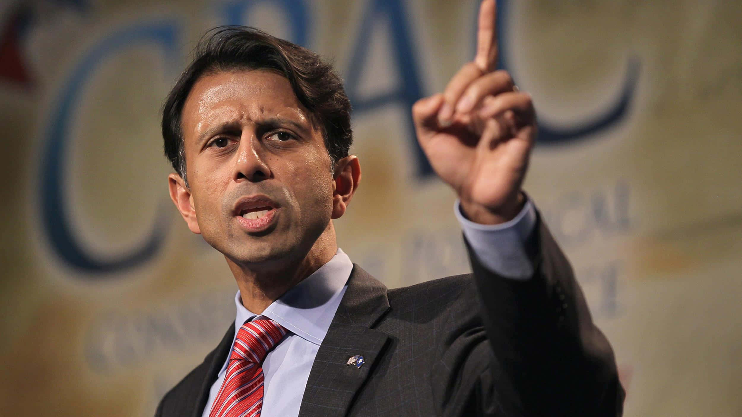 Bobby Jindal 在 CPAC 演講時指向上方 壁紙 — 一名身穿西裝、打紅色領帶的男子在模糊背景的政治活動中說話時，用力地做手勢。