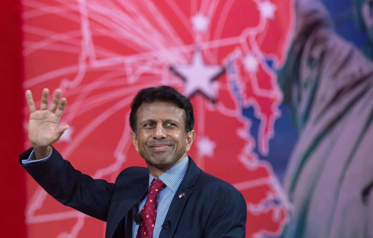 Bobby Jindal 在舞台上向觀眾揮手 壁紙 — 一個微笑的西裝男子在紅色背景（有白色星星和地圖輪廓）前向鏡頭揮手。