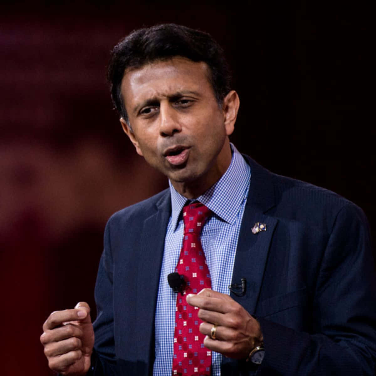 Bobby Jindal 在記者會上回答問題 壁紙 — 一名身穿深色西裝和紅色圖案領帶的男子在燈光昏暗的舞台上激情地做手勢演講。