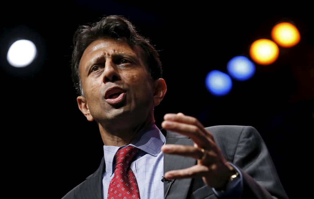 Bobby Jindal 表情生動的公開演講者 壁紙 — 一名身穿西裝、打紅色領帶的男子，在色彩繽紛的舞台燈光映襯的黑暗背景下，舉起手熱情地演講。