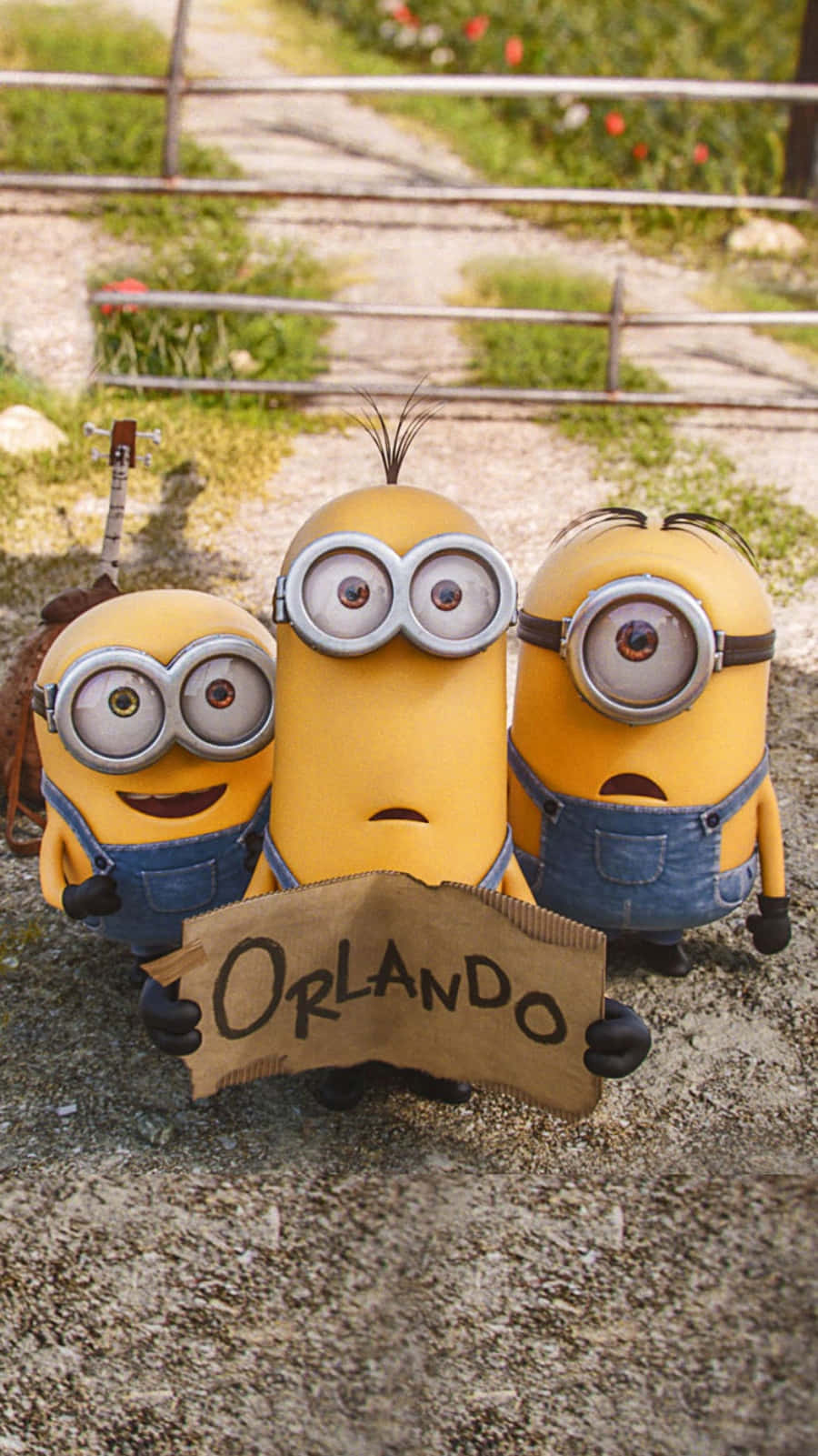 《神偷奶爸》Bob、Kevin 與 Carl 持「Orlando」紙條 iPhone 壁紙 — 三個穿著藍色工裝褲的黃色Minions在圍欄附近的碎石路上舉著寫有Orlando的紙板標語。