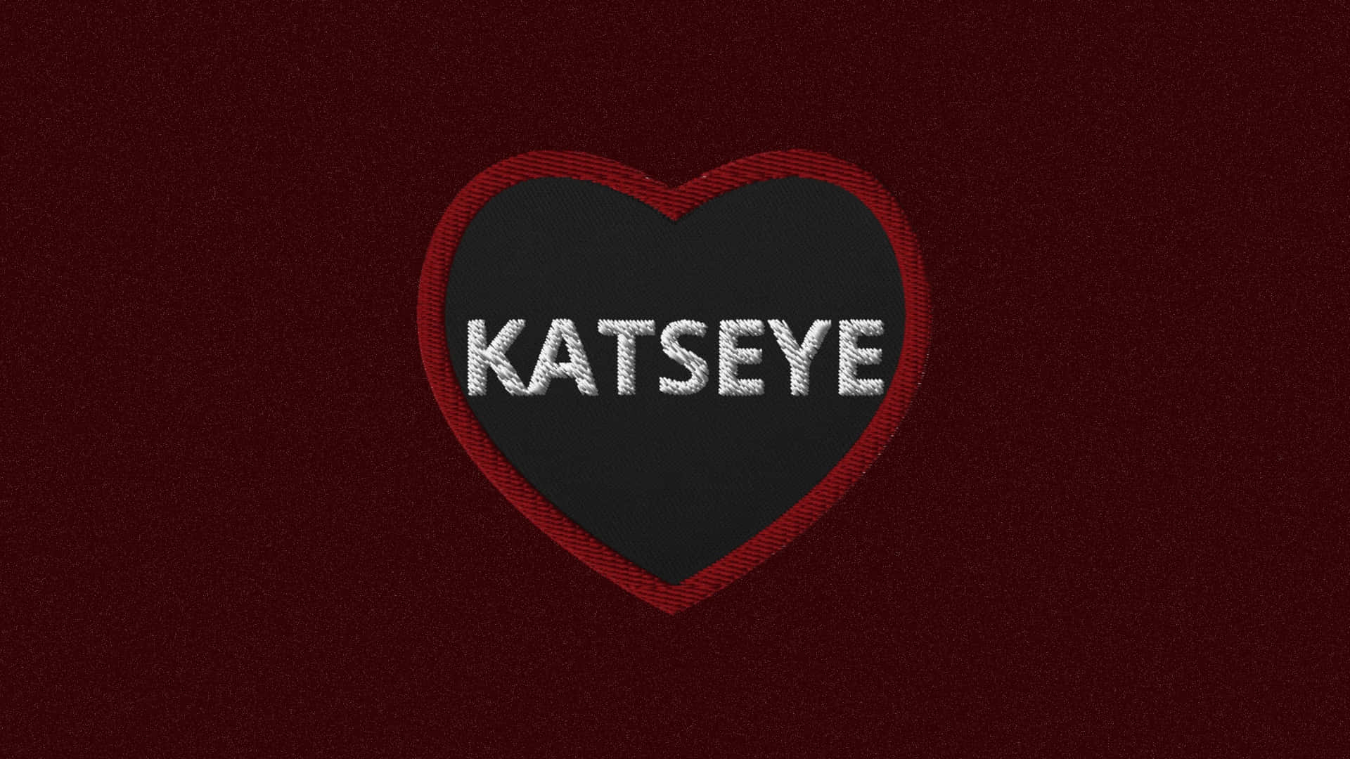  壁紙 — 一個大膽的刺繡心形補丁，中央以銀線繡有 'KATSEYE'，背景為深棗紅色布料。