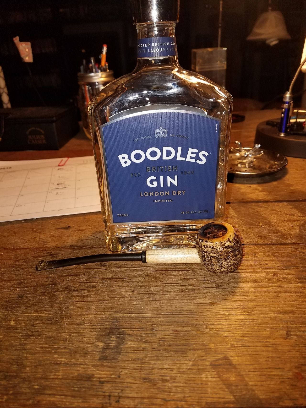 Boodles倫敦乾琴酒與煙斗 壁紙 — 一瓶 Boodles British Gin 和一個煙斗放在一個光線昏暗的房間裡的質樸木桌上。