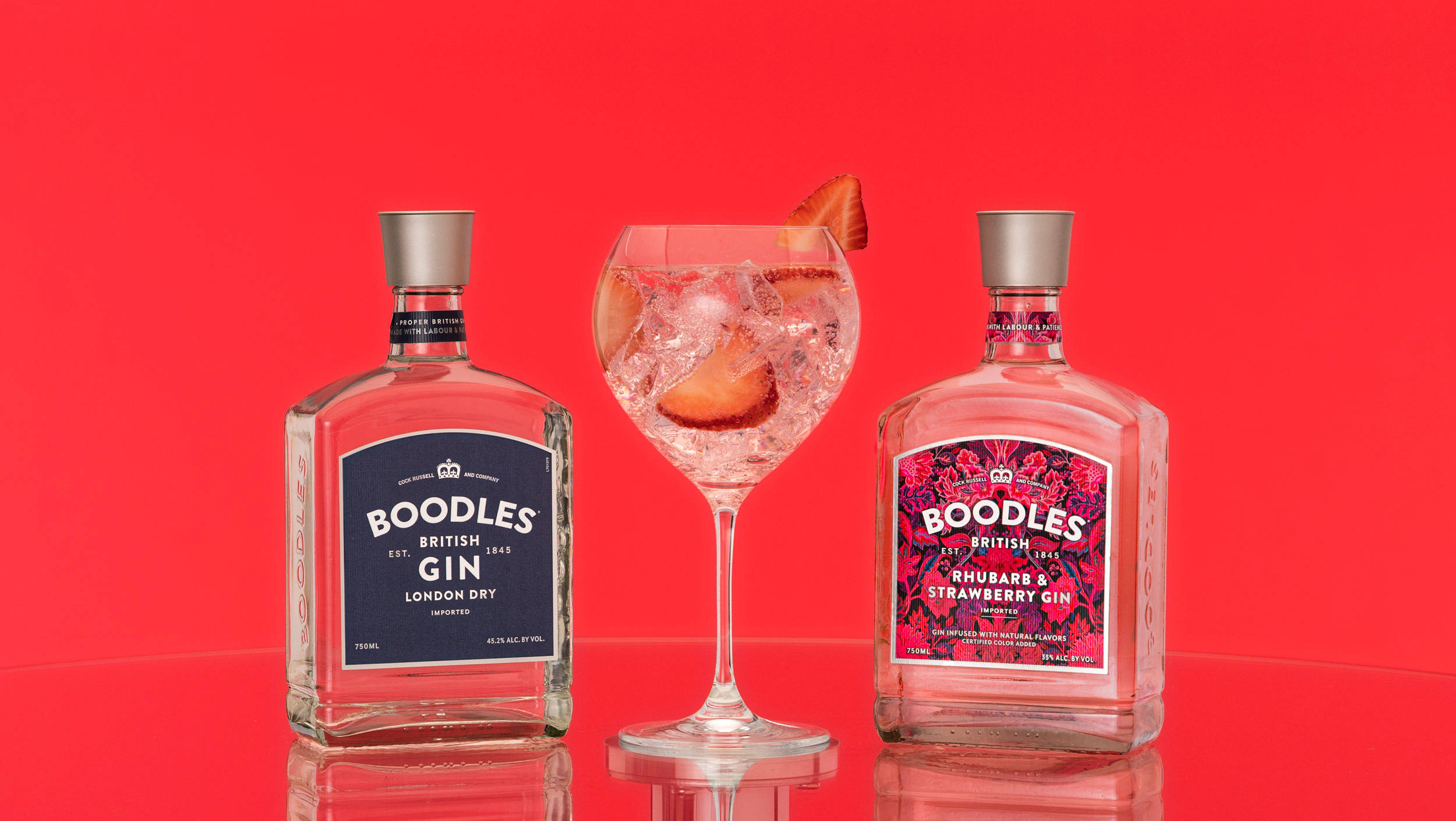 Boodles堅果與松針風味 壁紙 — 兩個Boodles氈酒瓶夾著一個裝滿冰塊和水果的氈酒雞尾酒杯，放在光滑的紅色表面上。