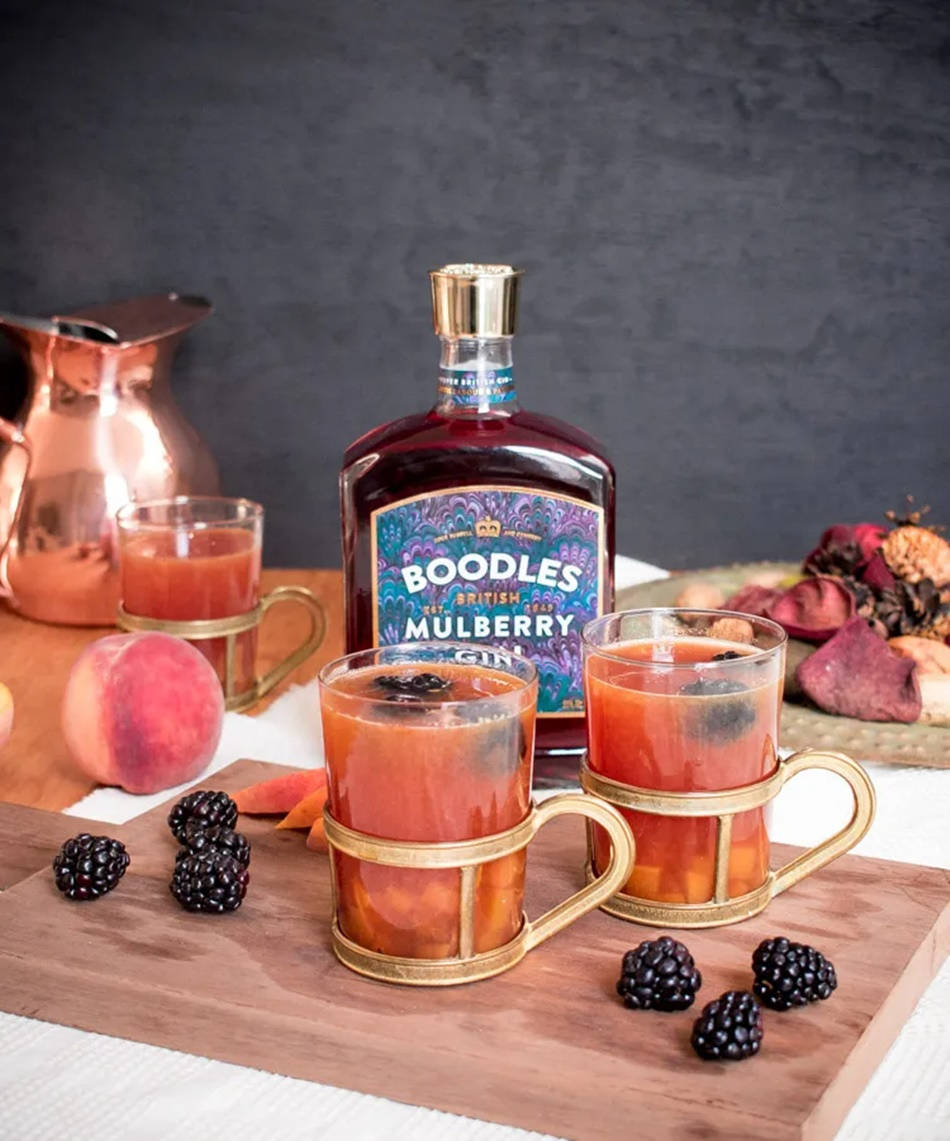 Boodles琴酒與莓果雞尾酒 壁紙 — 一瓶Boodles Mulberry Gin旁邊有兩個金色杯子，裝滿了莓果浸泡的飲料，放在木板上。
