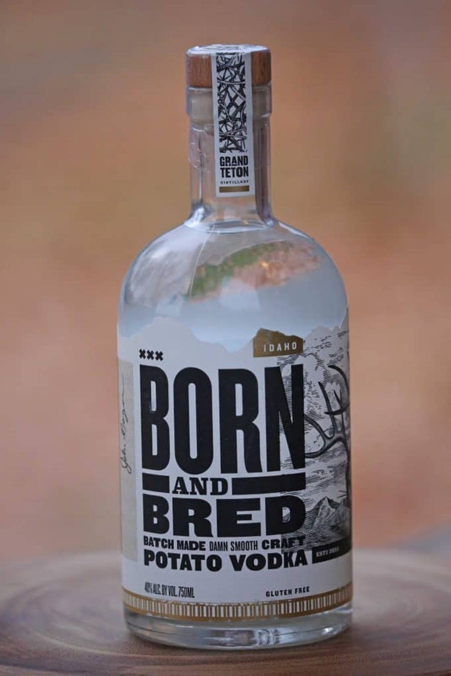 Born And Bred伏特加肖像照 壁紙 — 一個透明玻璃瓶的 Born and Bred 馬鈴薯伏特加，置於木質表面上，背景溫暖模糊。
