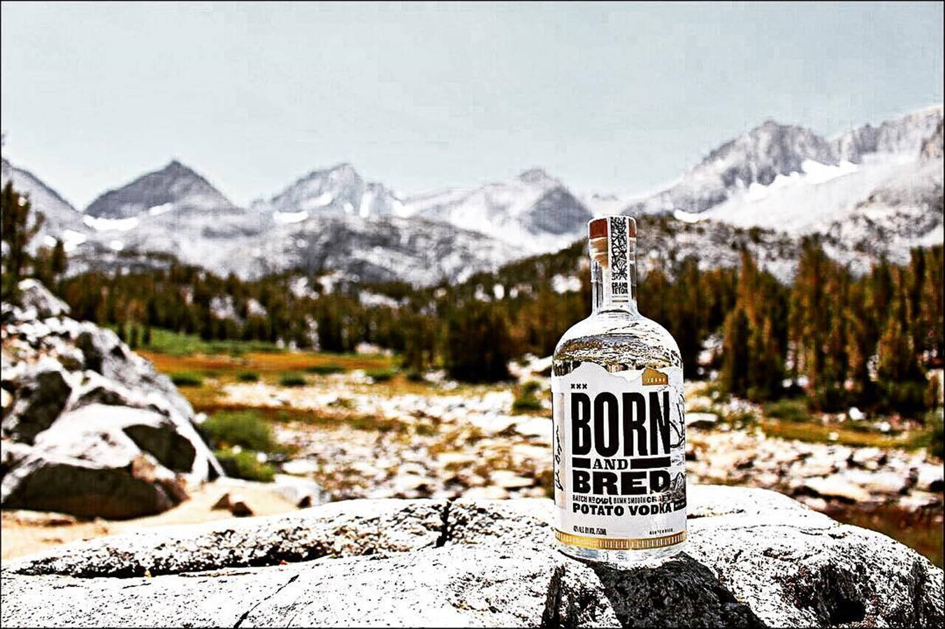 Born And Bred山景背景 壁紙 — 一瓶 Born and Bred 馬鈴薯伏特加放在雪山上嘅岩石上，背景係白雪覆蓋嘅山峰同常青樹。