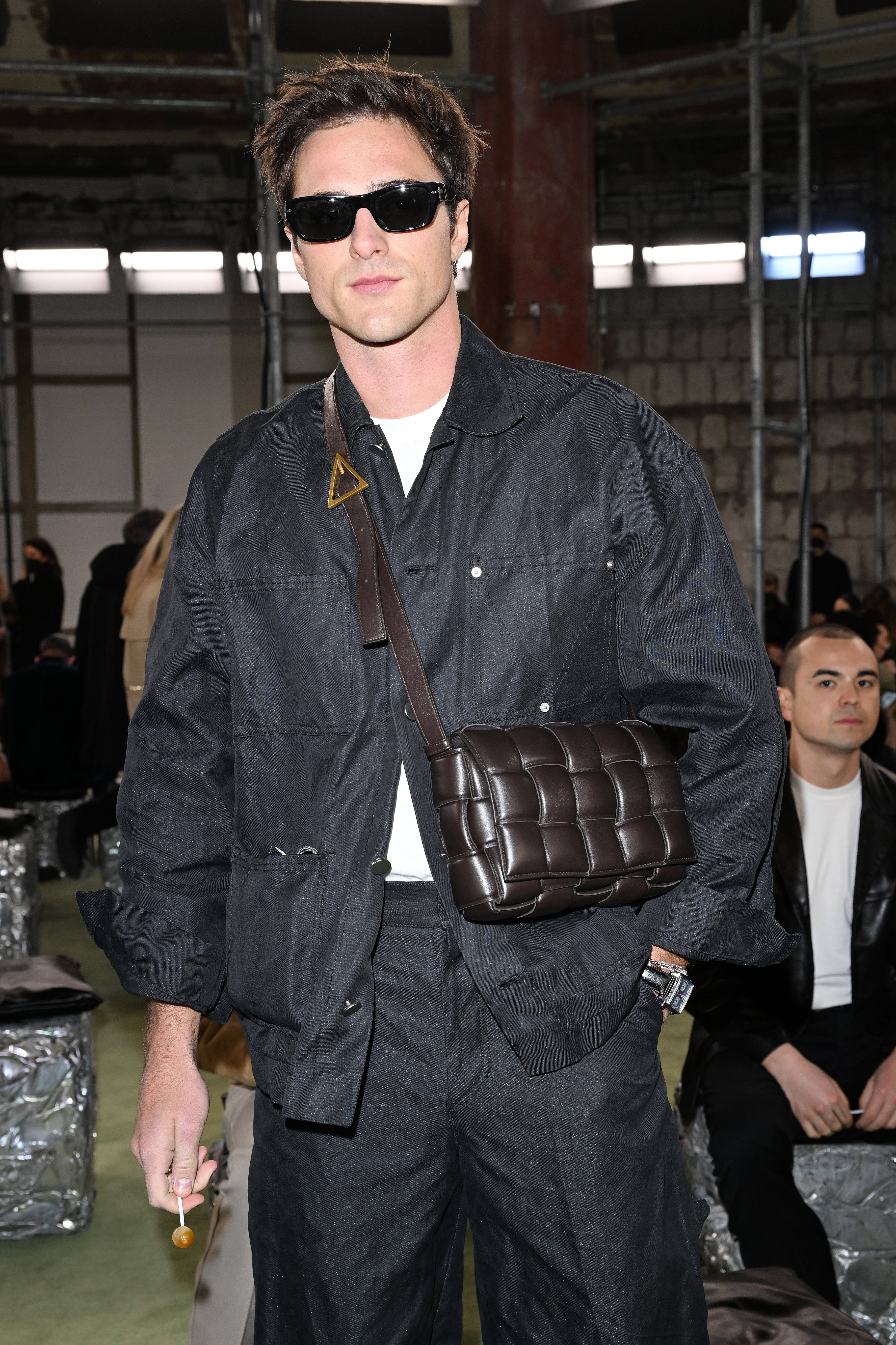 澳洲演員雅各布·艾洛迪展示 Bottega Veneta 手袋 壁紙 — 一名身穿深色工作服和太陽眼鏡的男子，在工業風格的時裝天橋場景中，手持一個絎縫皮革手袋，同時叼著香煙。