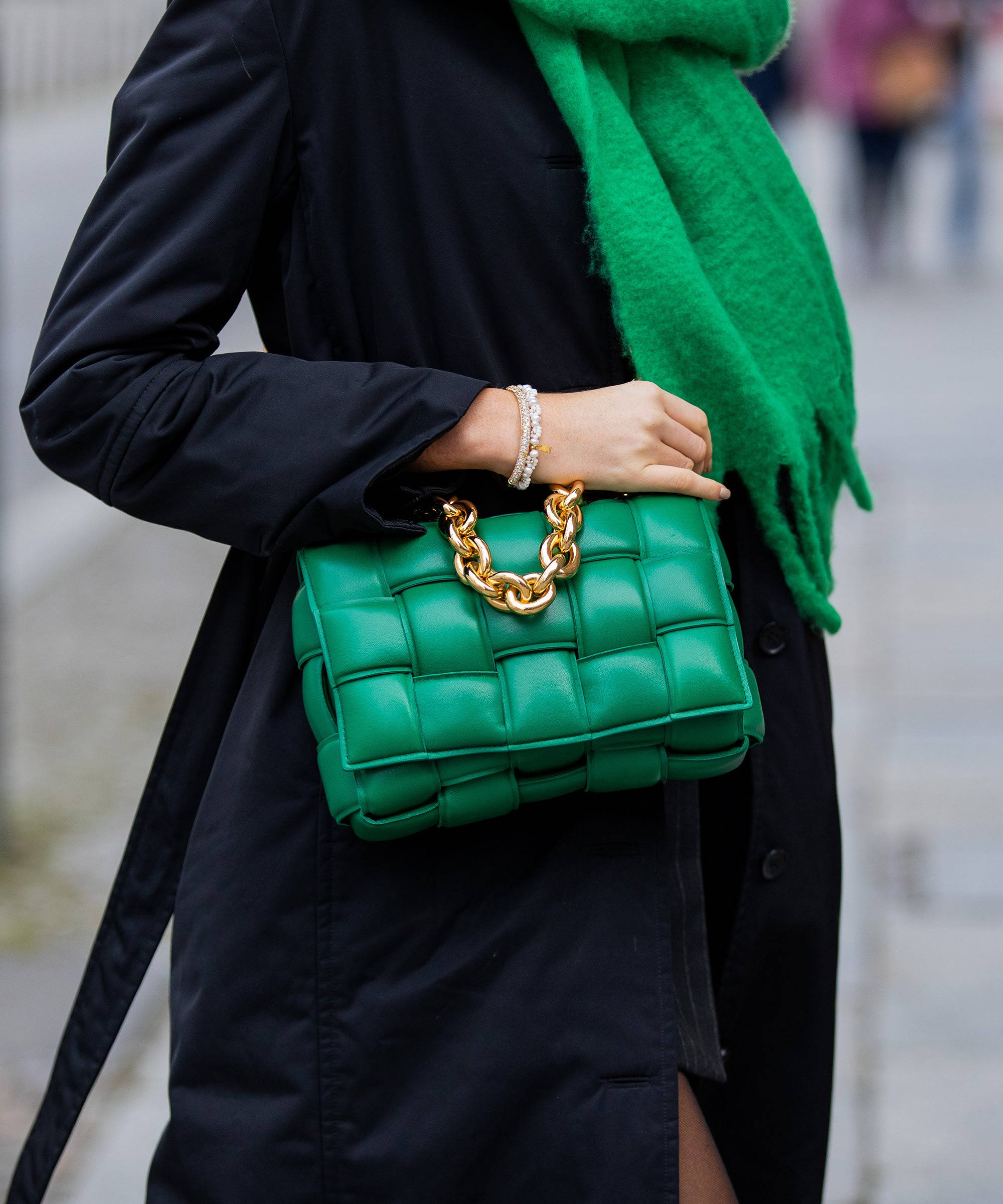 綠色 Bottega Veneta 手袋 壁紙 — 一名身穿黑色外套的人，在模糊的城市背景下，手持一個帶金色鏈條肩帶的鮮綠色編織手袋。