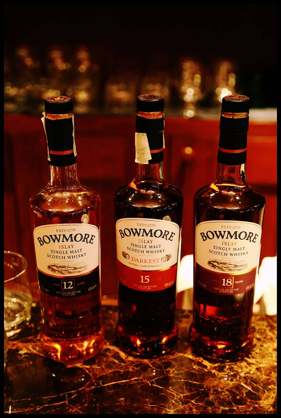 三瓶Bowmore威士忌在大理石吧檯上，溫暖燈光，背景有模糊的玻璃杯。