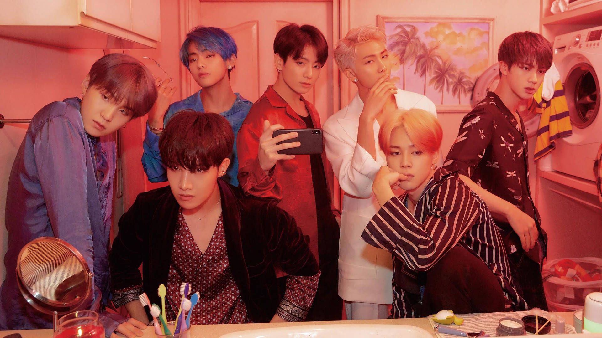 《Boy With Luv》粉紅氛圍BTS筆電壁紙 壁紙 — 七位擁有繽紛髮色的年輕男子在粉紅色燈光的浴室中擺姿勢，周圍是洗漱用品和一幅熱帶畫作。