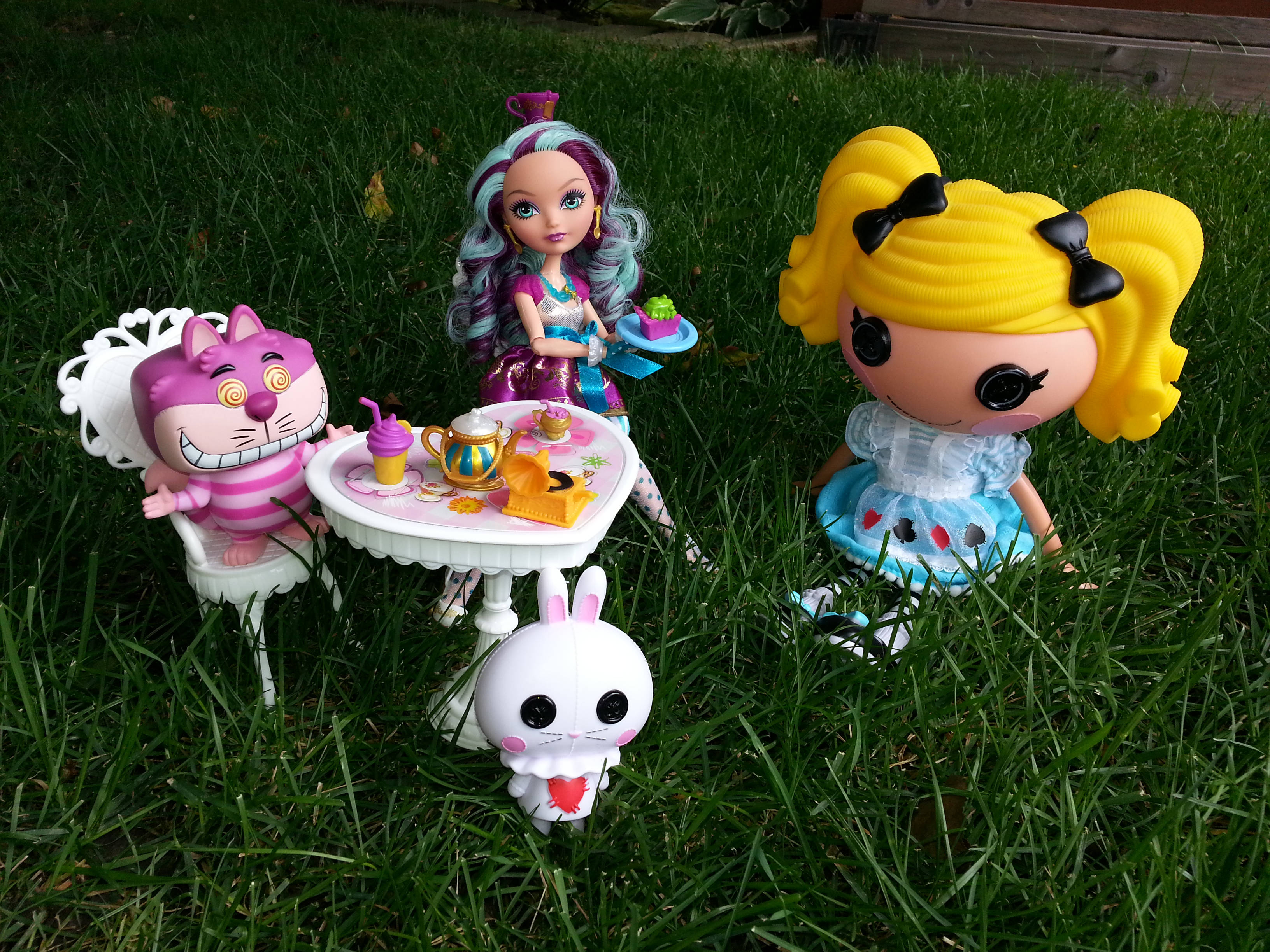 Bratz風格園林野餐 壁紙 — 三個風格化的玩具公仔和一個貓公仔聚集在一個微型茶具周圍，背景是充滿活力的綠色草地。