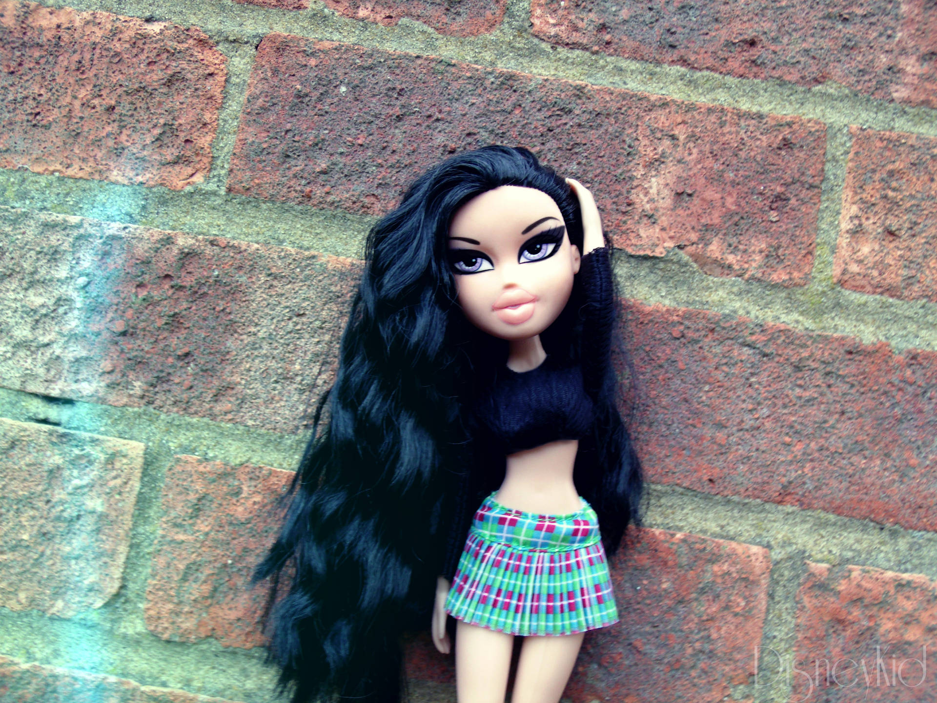 穿著格子裙的 Bratz 公仔靠在帶有綠色塗鴉的磚牆上。
