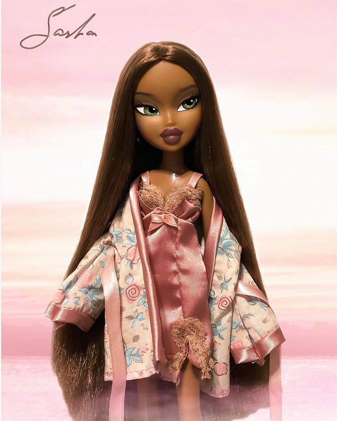 Bratz娃娃Sasha穿花袍 壁紙 — 一個擁有長長棕色頭髮和綠色眼睛的 Bratz 公仔，身穿粉紅色花卉睡袍，內搭緞面上衣，背景是柔和的粉紅色漸變。