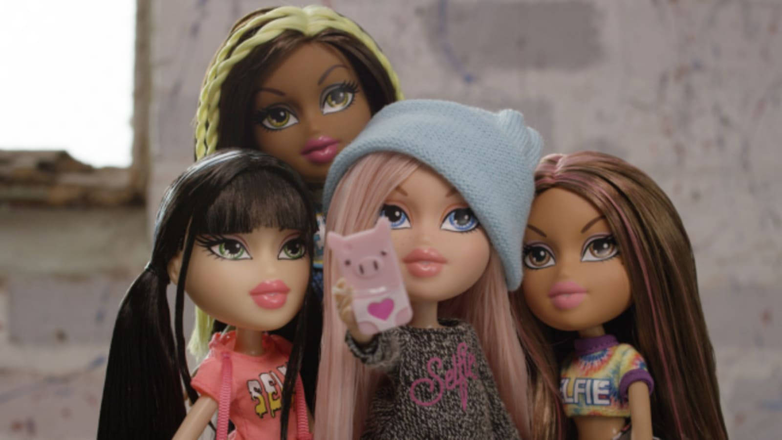 Bratz娃娃自拍 壁紙 — 四個 Bratz 公仔一齊擺甫士，其中一個喺風化嘅磚牆前面拎住一個豬主題嘅電話。