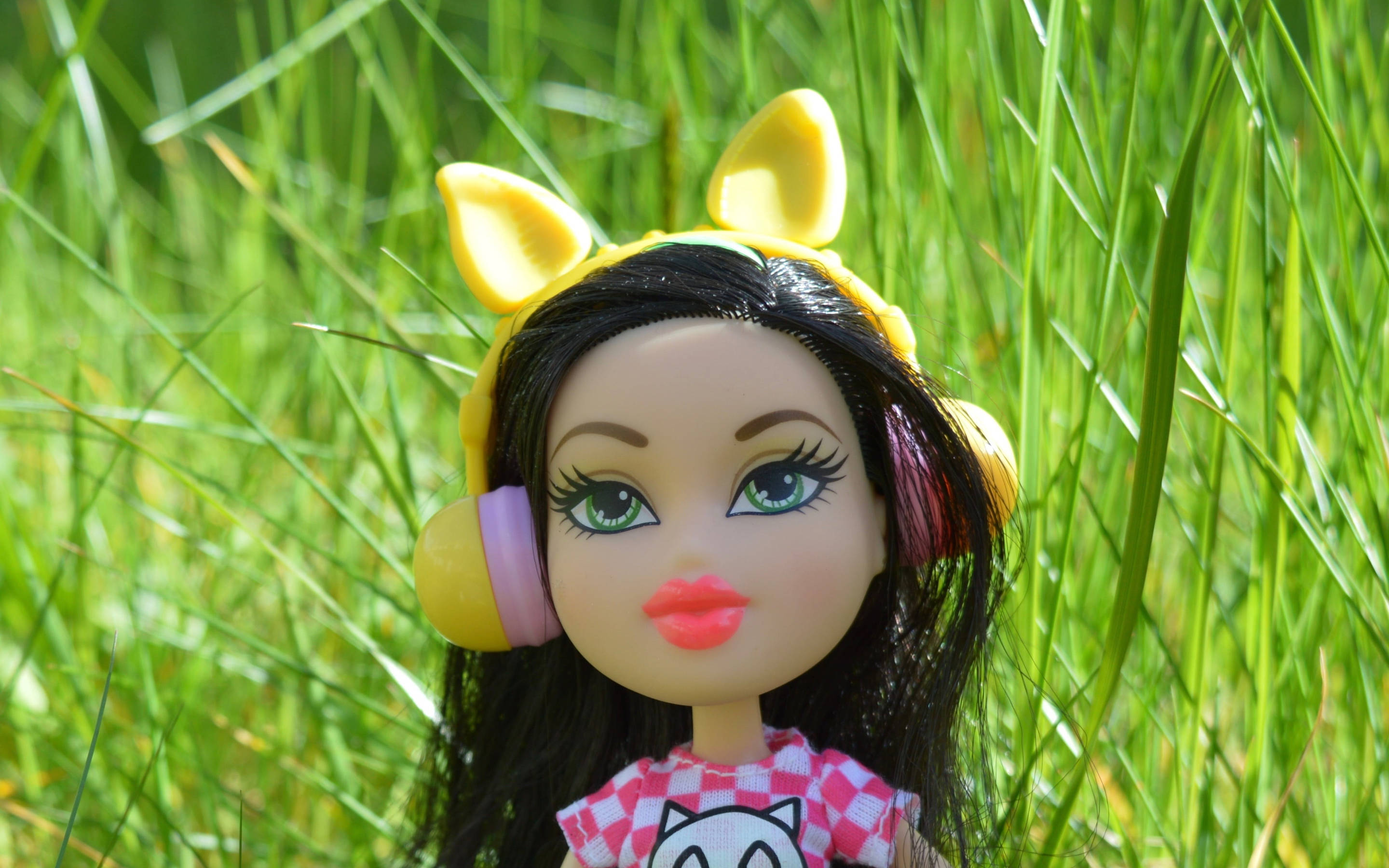 Bratz玩偶在花園 壁紙 — 一個戴著黃色貓耳和耳機的洋娃娃坐在陽光下明亮的綠色草地上。