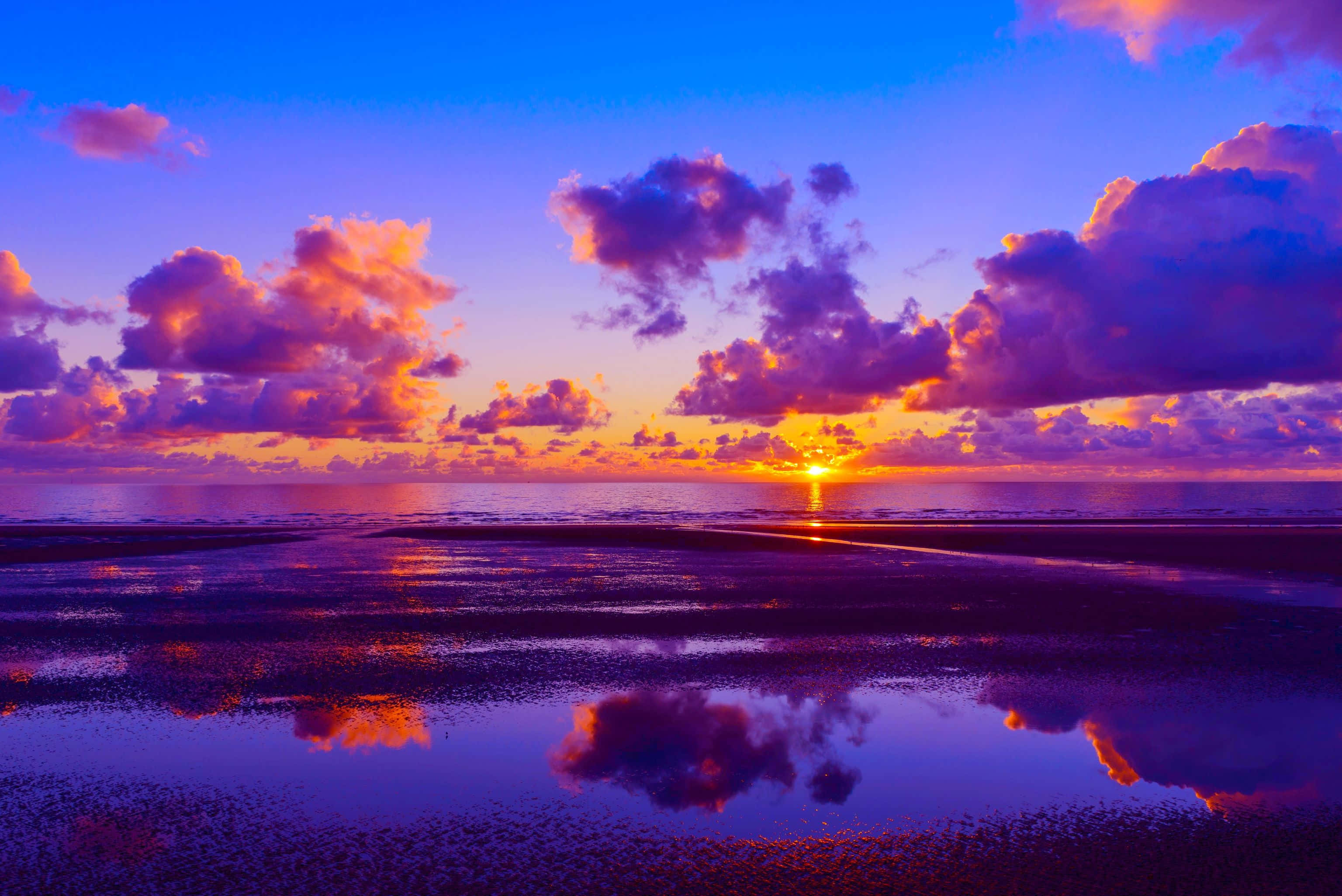 Colorful Sunset 桌布 壁紙 — 濕潤沙灘上令人驚嘆的日落，鮮豔的紫色、橙色和藍色雲彩倒映在淺水中。