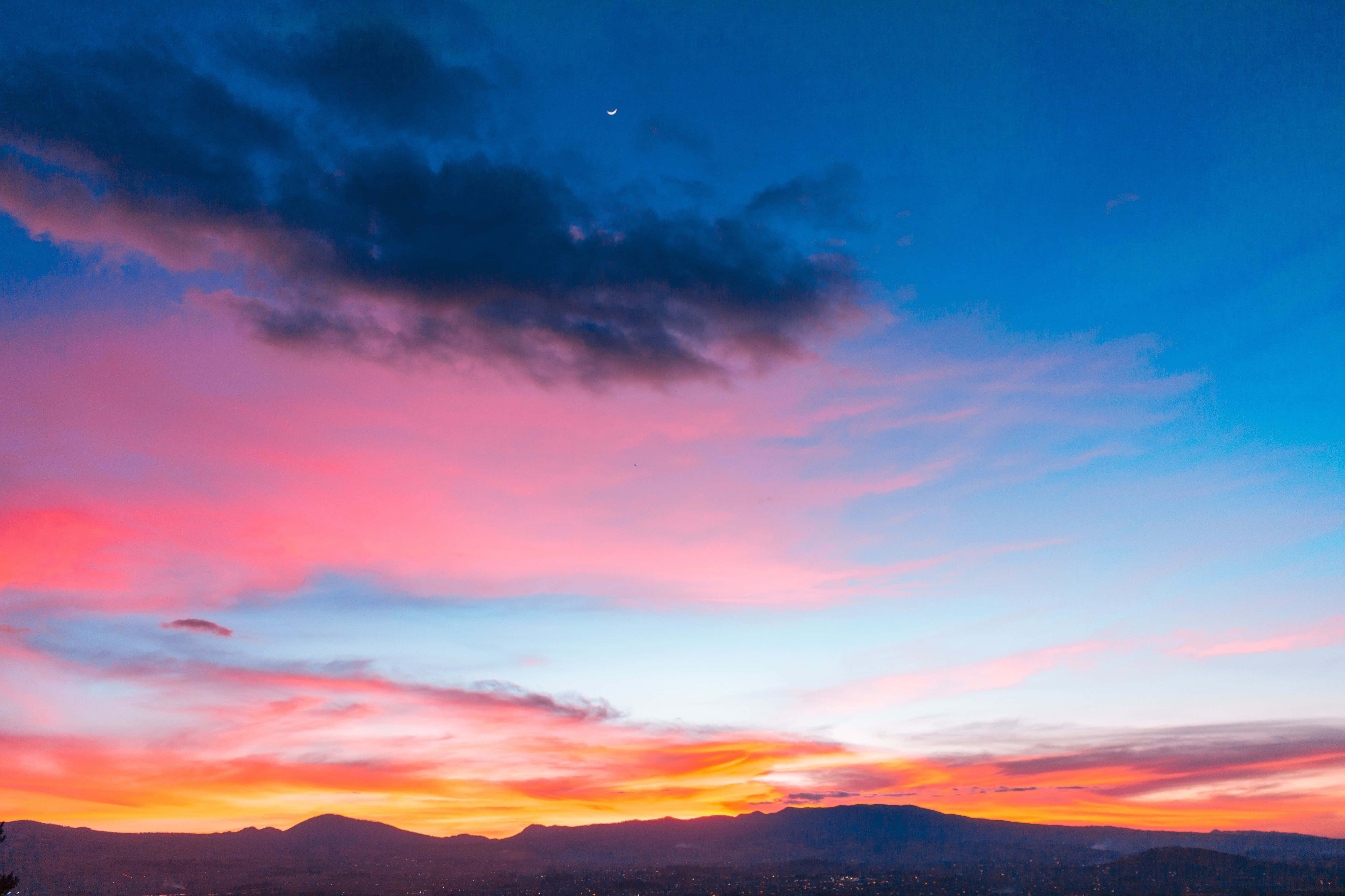 Colorful Sunset 桌布 壁紙 — 令人驚嘆的日落將天空染成火熱的橙色、粉紅色和深藍色，映照在遠山之上，可見一彎新月。