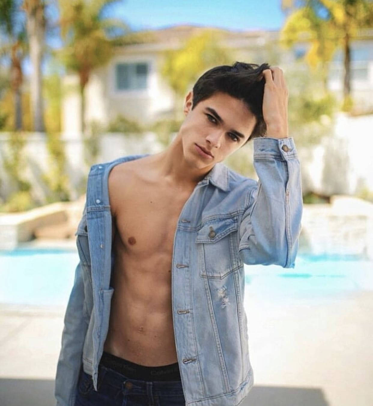 Brent Rivera 池畔聚焦照 壁紙 — 一位赤裸上身的年輕男子穿著牛仔外套，在陽光普照的泳池邊用手撥弄頭髮，身後有棕櫚樹和房子，背景柔和。