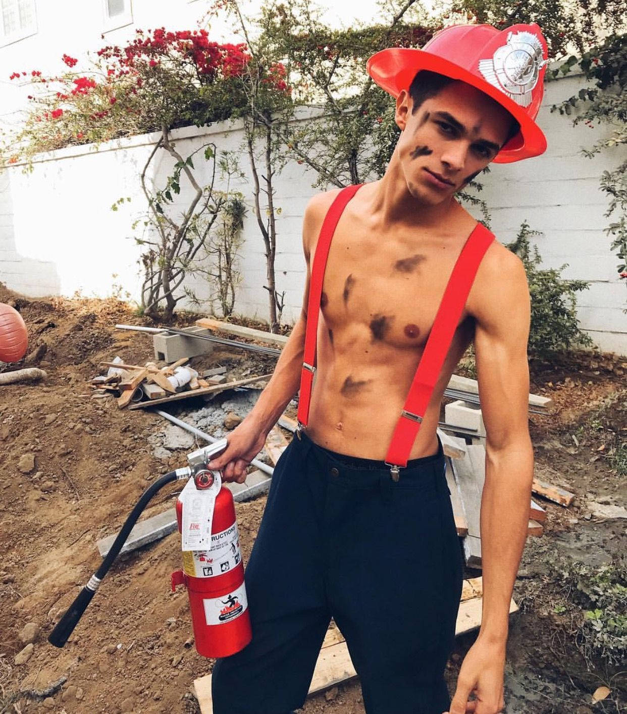 Brent Rivera 穿消防員服裝 壁紙 — 一個赤膊的年輕男子穿著紅色吊帶和消防員帽，在雜亂的戶外建築工地中拿著滅火器。
