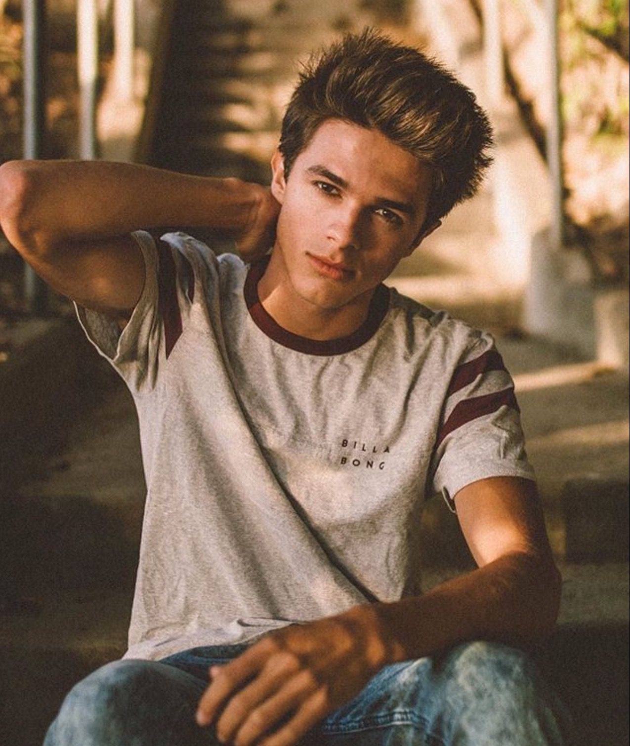 Brent Rivera 美學風格照 壁紙 — 一位年輕男子頭髮造型時尚，穿著灰色Billabong T恤和牛仔褲，在溫暖的金色陽光下隨意坐在階梯上。