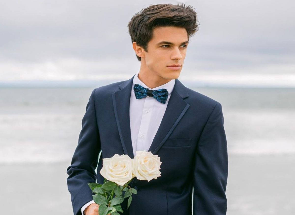 Brent Rivera 西裝典雅照 壁紙 — 一名身穿海軍藍西裝和圖案領結的年輕男子在多雲的海灘上手持兩朵白玫瑰。