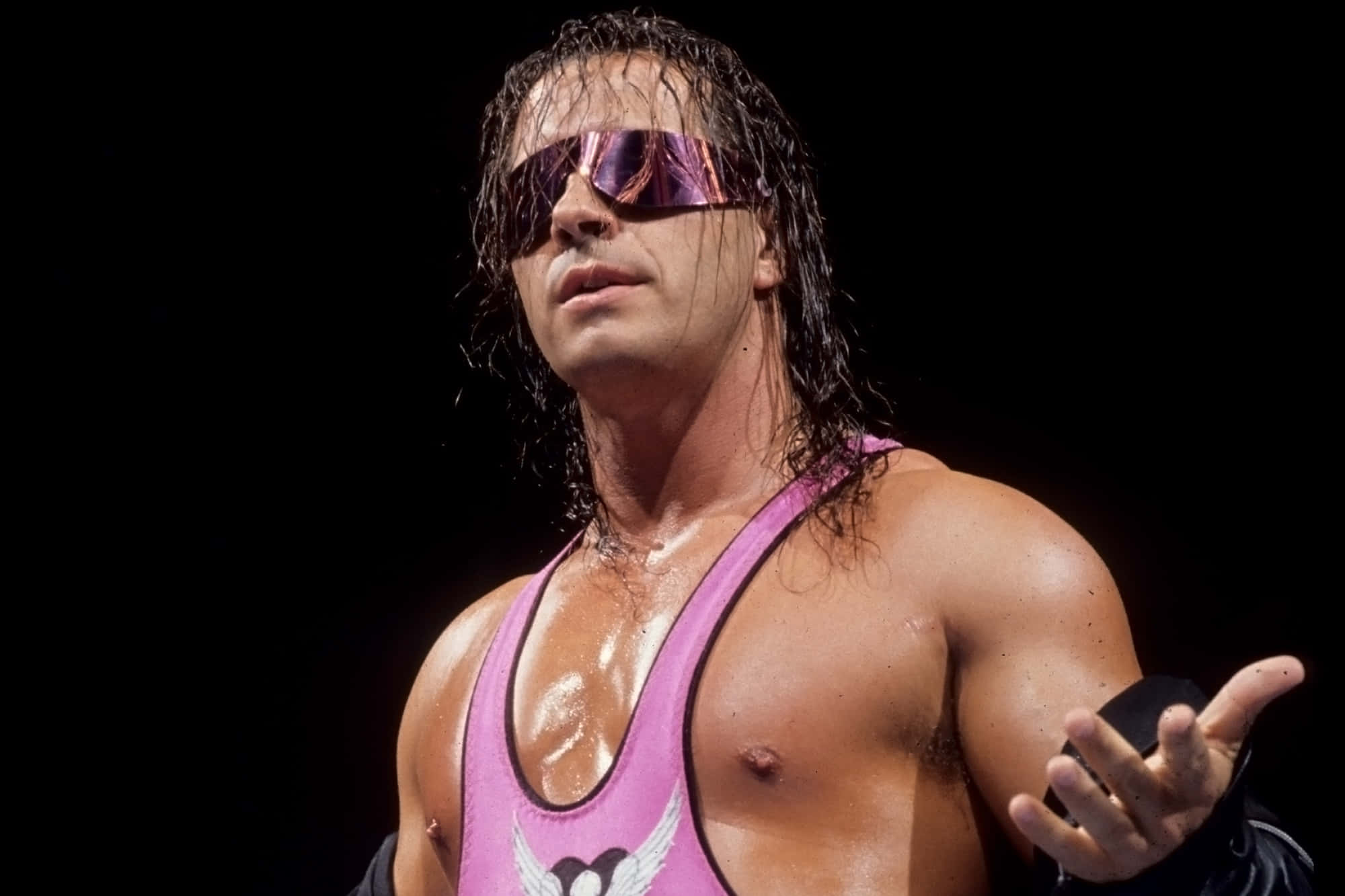 Bret Hart WWE帥氣摔角手 壁紙 — 一位肌肉發達的摔角手穿著粉紅色緊身衣和反光太陽眼鏡，濕髮，張開手掌，自信地站在黑色背景前。