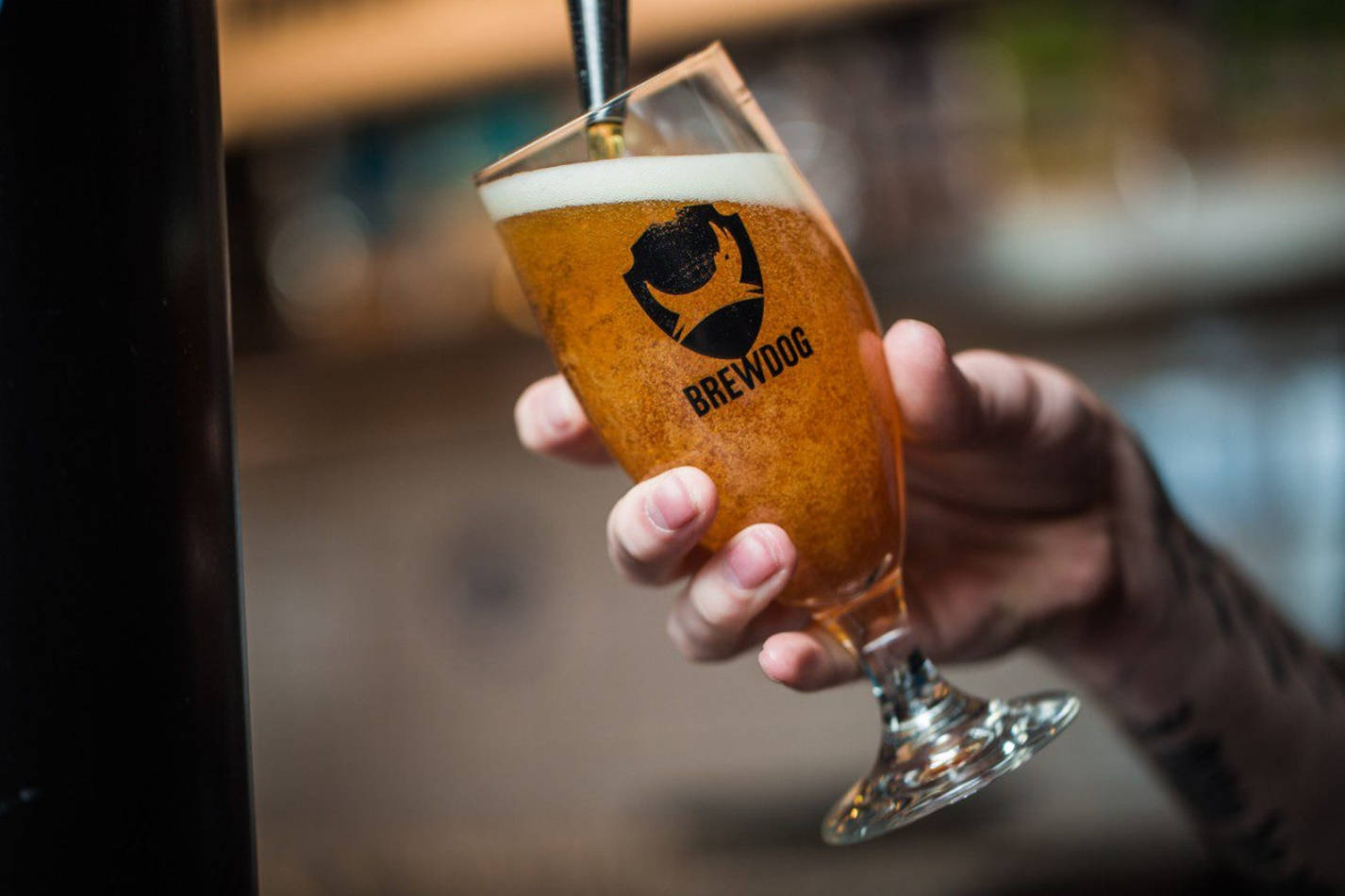 BrewDog 啤酒倒入酒杯 壁紙 — 一隻手拿著一杯琥珀色啤酒，帶有白色泡沫，從酒吧的水龍頭倒出。