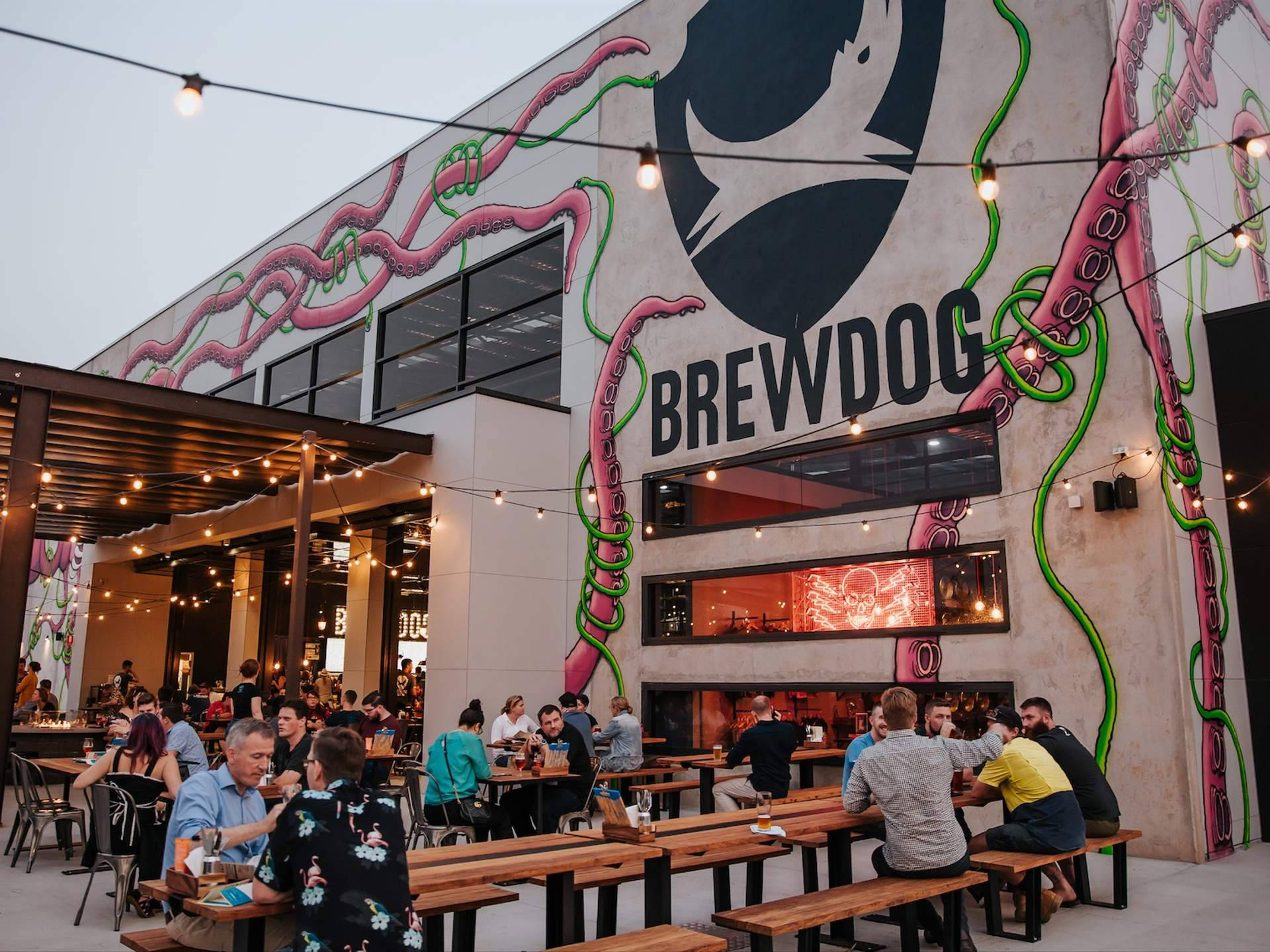 BrewDog DogTap 布里斯本外觀 壁紙 — 人們在戶外餐桌用餐，上方有燈串，背景建築物有鮮豔的粉紅色和綠色八爪魚壁畫以及大型BrewDog標誌。